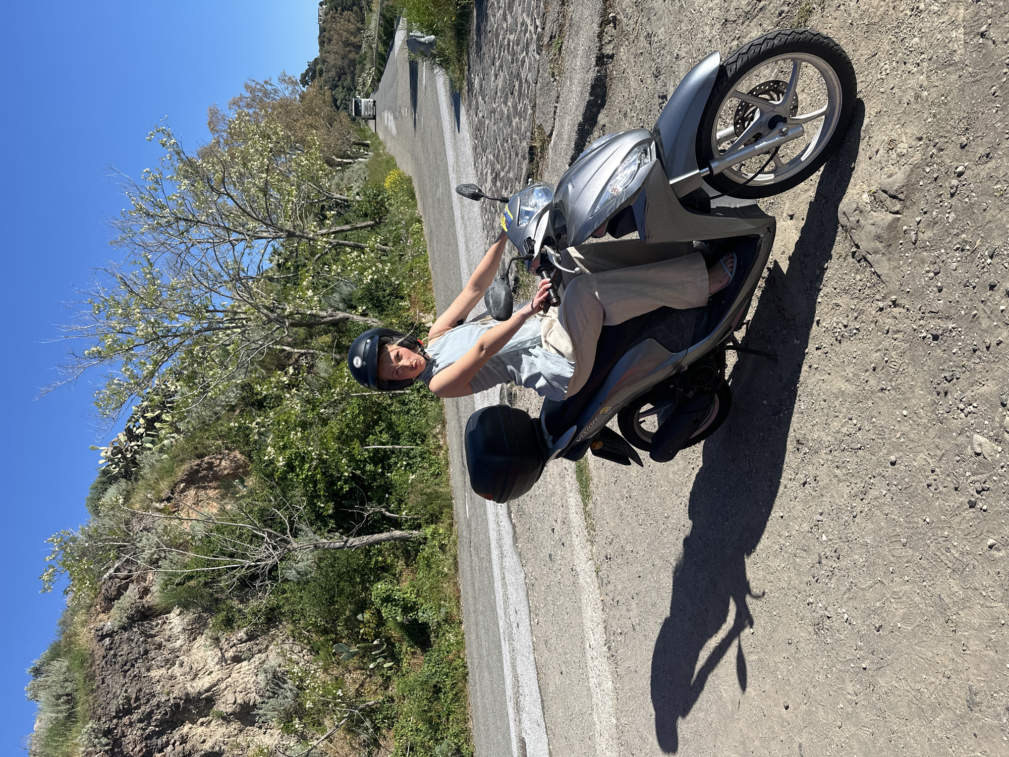 The image features a woman seated on a silver scooter on a sunny day, likely in Lipari, Italy. She is wearing a black helmet, a light blue t-shirt, light-colored pants, and sandals, and is holding the handlebars. The scooter is a Honda Vision model, with its name visible on the side panel. A black storage box is attached to the rear of the scooter.
The scene is an outdoor setting on a narrow, winding road that appears to be a mix of paved and dirt/gravel surfaces. To the left, a steep hillside is covered with abundant Mediterranean vegetation, including trees with white blossoms, green bushes, and various cacti and succulents. The sky above is a clear, brilliant blue. Strong, distinct shadows from the scooter and the woman are cast on the ground, indicating bright midday or early afternoon sun. In the distance, further up the road, a white vehicle is visible. The overall impression is a scenic, warm weather location, suggesting a stop during a journey.
