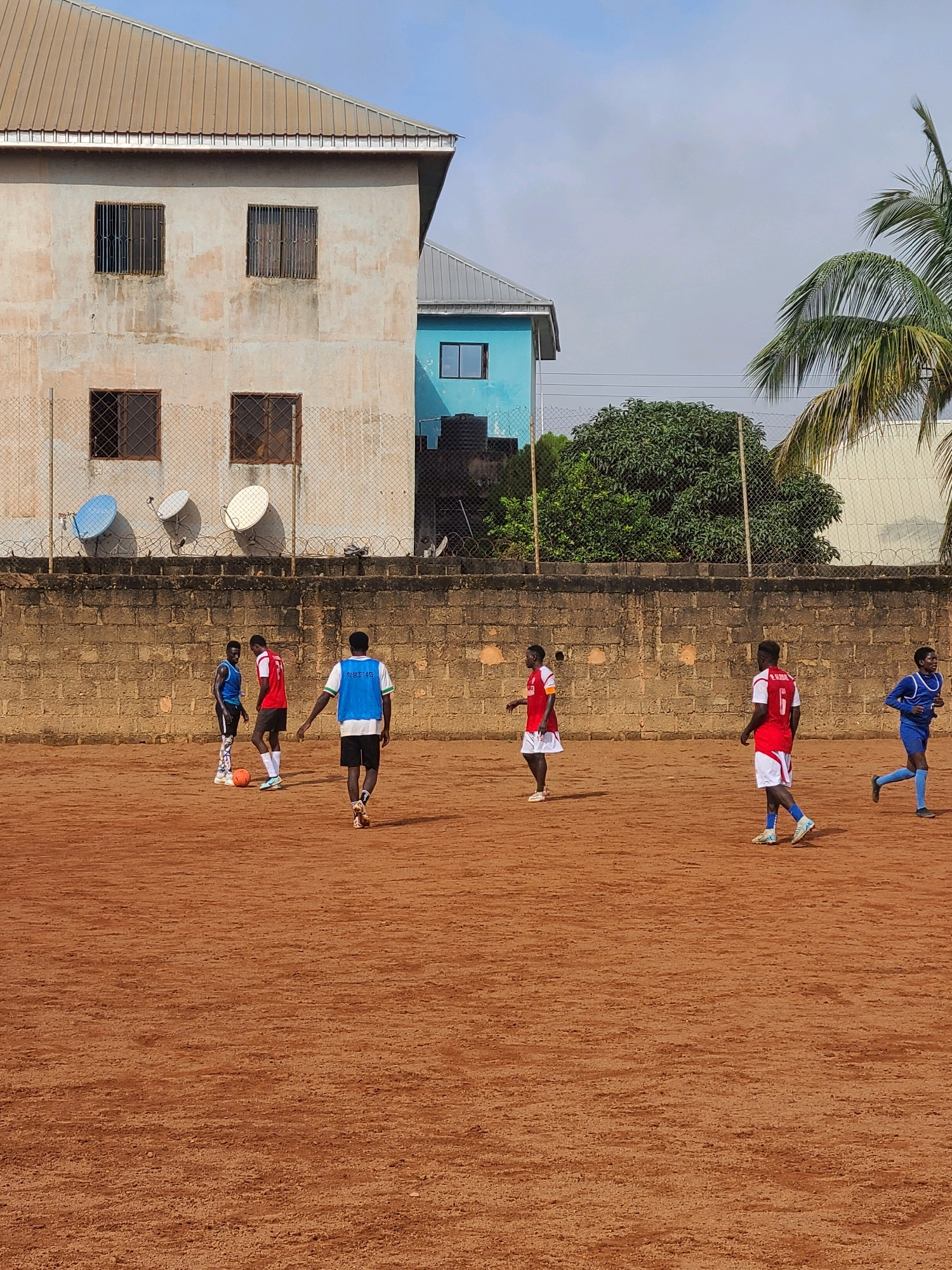 The image depicts an outdoor soccer game on a dirt field in Barakin Lerawi, Nigeria. Several young men are actively engaged in the sport, wearing red and white or blue uniforms. One player in the foreground, wearing a blue vest over a white shirt and black shorts, walks away from the camera, his back to the viewer. In the background, two buildings are visible: a light-colored, weathered two-story structure with barred windows, and a smaller, vibrant blue building. A brick wall runs along the perimeter of the field, behind which are some trees and a palm tree. The sky is overcast.