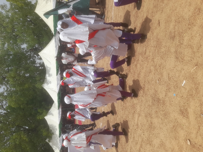 A group of individuals wearing white garments with red sashes and purple leggings stand in a line outdoors. They are positioned on sandy ground under a white canopy with green accents. Trees are visible in the background. The formation and attire suggest a ceremonial or performance context.