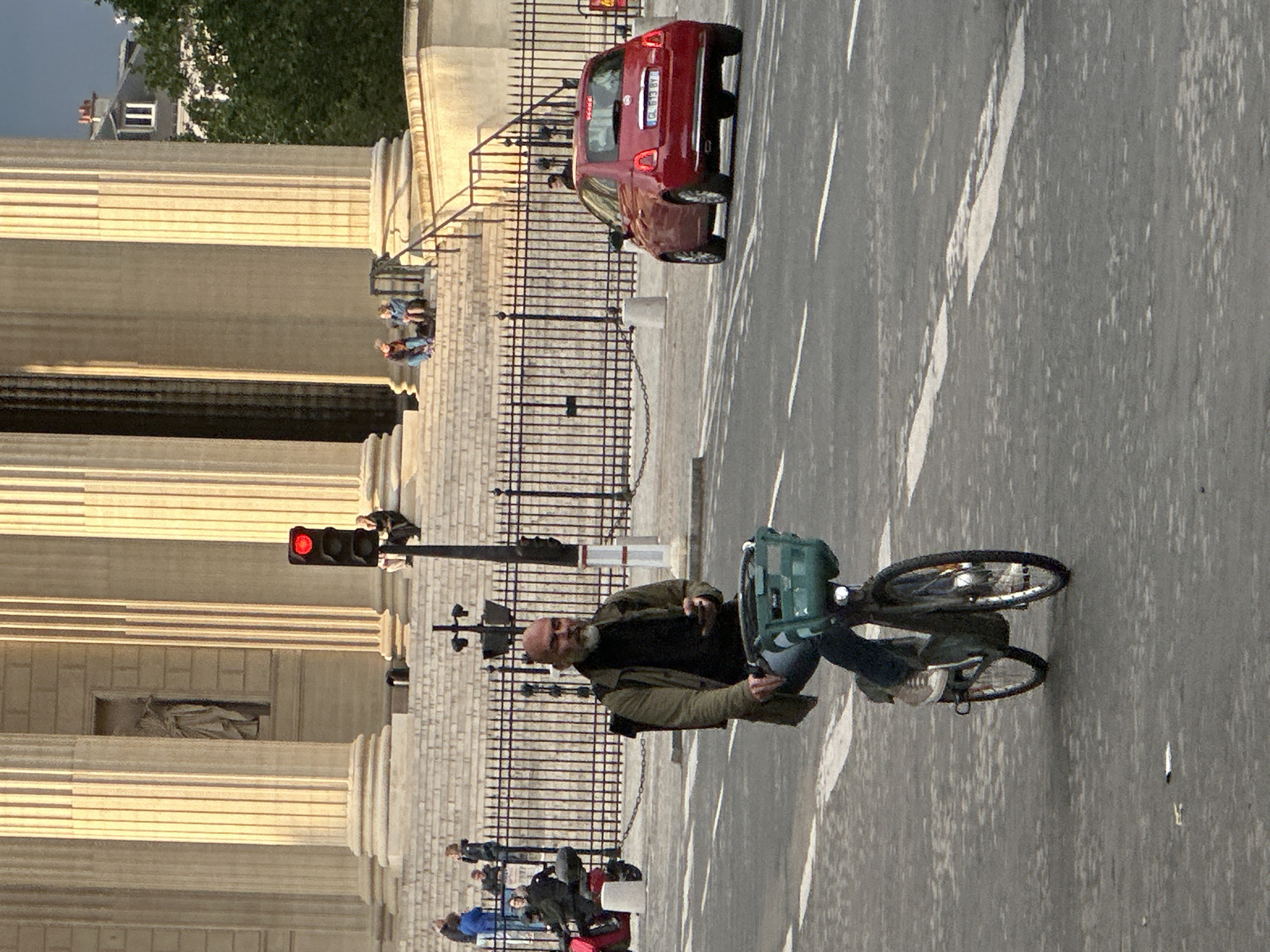 The image shows a street scene in Paris, France, with the Église de la Madeleine church visible on the left. A man rides a bicycle on the right side of the street. A red car is driving in the opposite direction. People are sitting on the steps of the church. The traffic light is red. A black wrought iron fence lines the sidewalk next to the church.