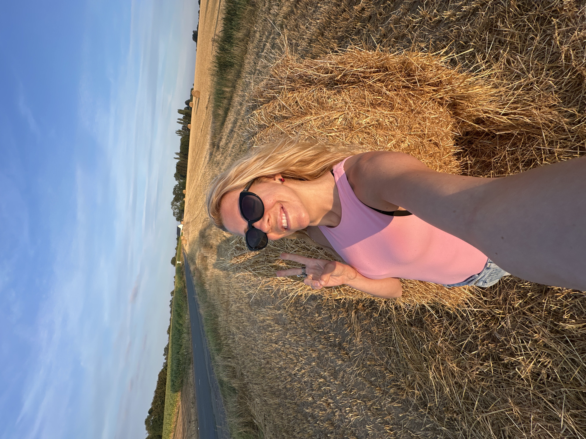 In a field of harvested crops, likely near Trouville-Alliquerville, France, a woman is taking a selfie. She has blonde hair and is wearing sunglasses and a pink tank top with denim shorts. She is smiling and holding up two fingers in a peace sign. Behind her, hay bales are stacked. The setting sun casts long shadows across the field, and the sky is a clear blue with a few wispy clouds. A paved road runs along the edge of the field in the background, with trees and more hay bales visible in the distance.