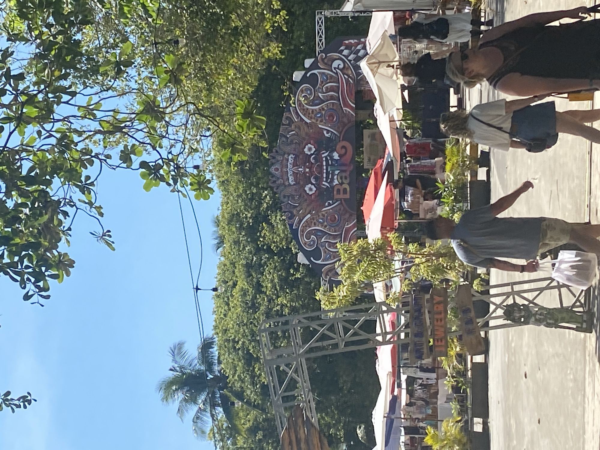 The image depicts a bustling outdoor market in Kuta Selatan, Indonesia. Numerous vendors are present with stalls sheltered by a mix of white, red, and blue umbrellas. A large sign decorated with a Balinese design, marked with "Bali," serves as a backdrop. People are walking through the market, some browsing goods and others passing by. Trees are visible on the left side of the image, with a blue sky overhead. A metal structure with signs for "Jewelry" is in the foreground on the right side of the image.