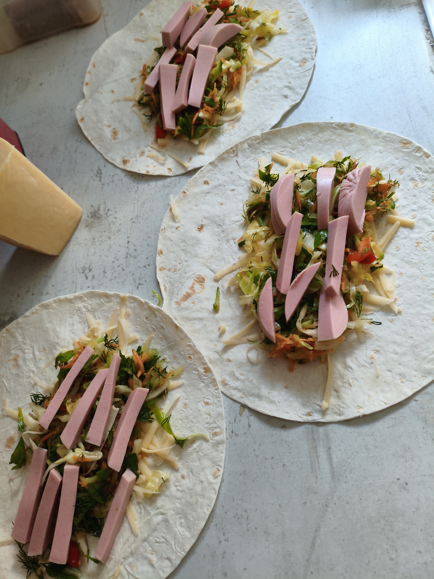 Three lavash breads are laid out on a gray, speckled surface, each topped with shredded cheese, a mix of chopped vegetables including cabbage, carrots, and peppers, and sliced pink sausages. A wedge of yellow cheese is visible in the lower left corner. The scene depicts food preparation, likely for shawarma or wraps. The image does not provide any information that could be used to determine its location in Orenburg, Russia.