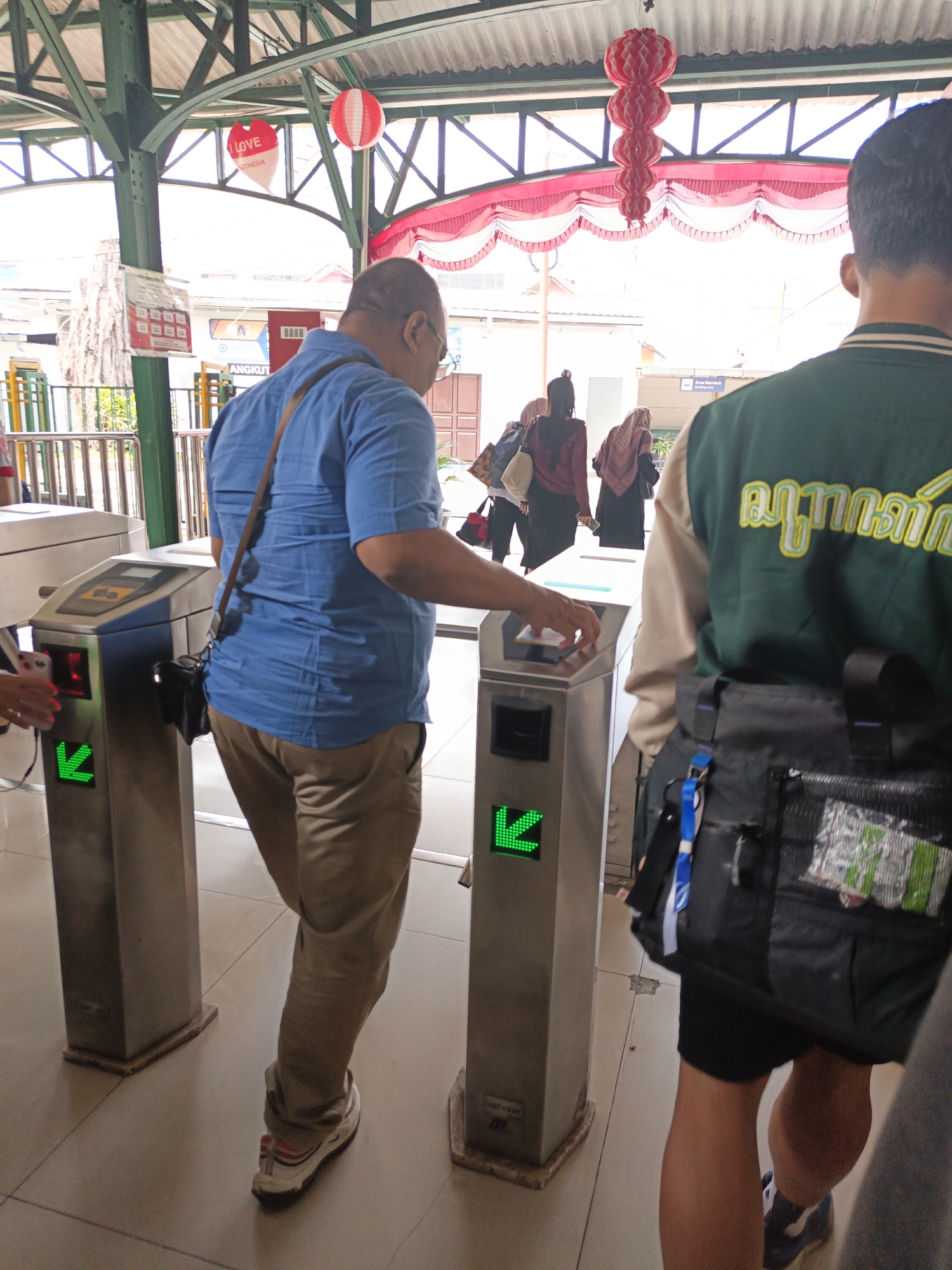 The image shows a man in a blue shirt and tan pants passing through an electronic gate at a train station in Surakarta, Indonesia. He is carrying a brown bag over his shoulder. Another person, partially visible, is walking behind him. In the background, several women wearing hijabs are also visible. Red and white decorations, possibly for a celebration, are hanging from the ceiling. The gate has a green arrow pointing left, indicating the direction of entry. The environment appears to be an indoor or covered area with a tiled floor.