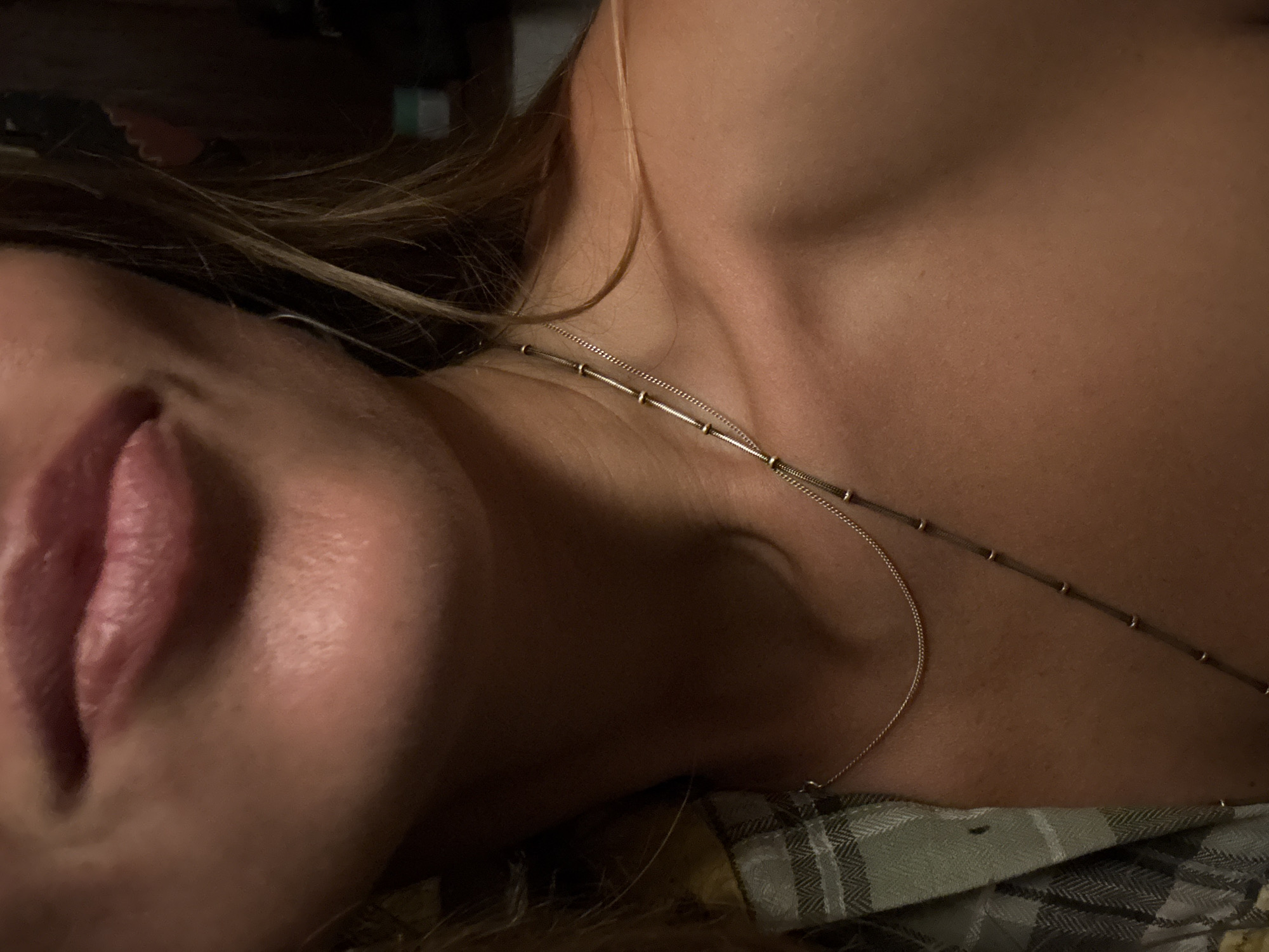 The image is a close-up, chest-up shot of a person's neck, shoulders, and mouth. The person has fair skin and light brown hair. They are wearing a necklace with a delicate chain and a longer chain with small beads. A portion of a green and gray plaid shirt is visible on the left side of the frame. The background is dark and out of focus, with no discernible city details. The lighting is dim and appears to be artificial.