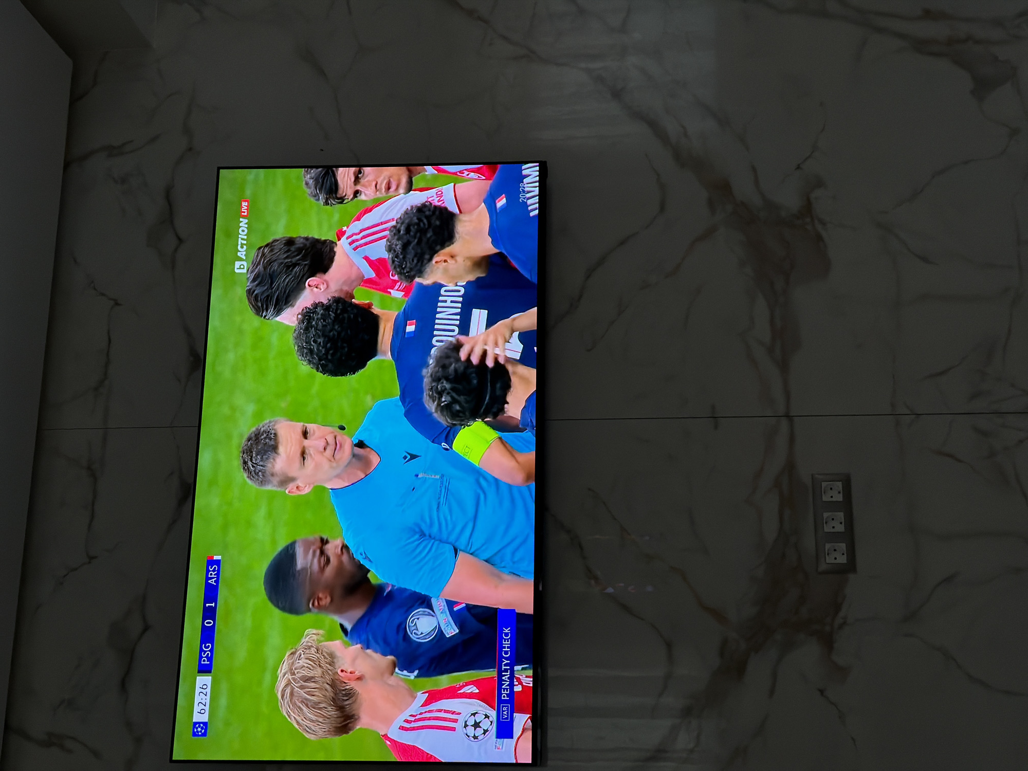 The image shows a television screen displaying a soccer match. The score is PSG 0 - 1 ARS (Arsenal). The time on the clock is 62:26. The bottom of the screen indicates "VAR PENALTY CHECK". The players are wearing soccer jerseys and the referee is wearing a blue shirt. The background behind the TV is a marble-patterned wall with three electrical sockets visible. The image does not contain sufficient information to identify its location in Asenovgrad, Bulgaria.