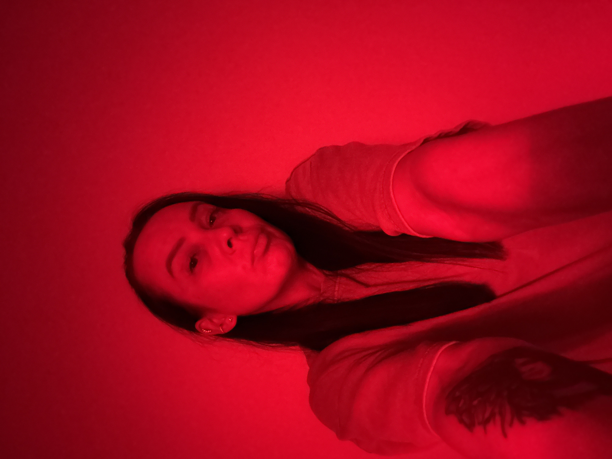 A single adult female is depicted in a medium-shot selfie, illuminated entirely by a monochromatic red light, ostensibly within a private indoor setting in Chaikovsky, Russia. The subject, possessing long dark hair, gazes directly at the camera with a slight head tilt and pursed lips. She wears a light-colored, long-sleeved garment rendered red by the pervasive lighting. Multiple metallic earrings are visible on her left ear. A dark, possibly abstract or floral, tattoo is present on her right inner bicep/forearm. The background is a plain, undifferentiated wall, uniformly red due to the lighting. The image is captured from a low angle, suggesting the subject holds the camera at arm's length.