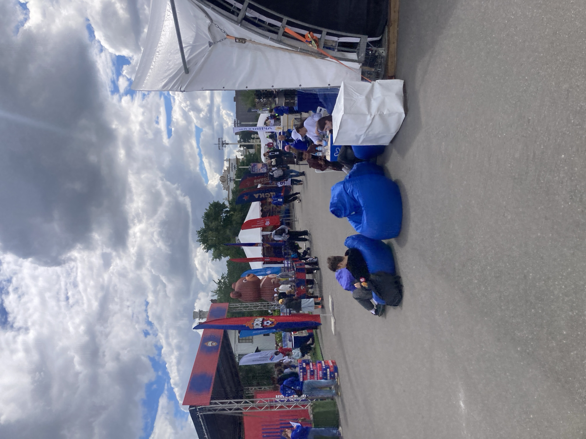 The image captures an event taking place in Moscow, Russia. The scene includes a variety of elements: tents, tables staffed with people, flags, and a large inflatable bear. People are milling about. The presence of flags bearing the logo of the Russian football club CSKA Moscow suggests a connection to the team. The architecture in the background points towards a location within Moscow, Russia. A child is near some blue inflatable chairs.