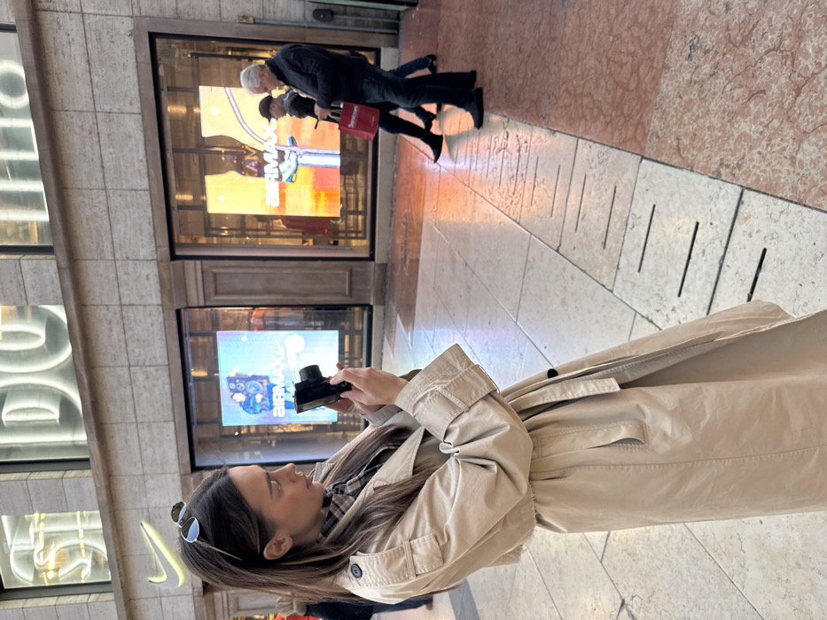 An outdoor scene is captured on Via Giuseppe Mazzini in Verona, Italy, in front of a Nike retail establishment. In the foreground, a young adult female, with long brown hair and sunglasses resting on her head, stands in profile facing right. She is attired in a light beige trench coat over a plaid shirt and is holding a black compact digital camera with both hands, aiming it towards an unseen subject to her right. The background features the Nike store's light-colored stone facade, incorporating large display windows with bright digital screens exhibiting "AIRMAX" branding and product imagery. Above the storefront, large illuminated letters spelling "NIKE" and "DO" (partially visible) are affixed. In the mid-ground, three individuals are walking from right to left. The leading figure is an older male with grey hair, dressed in a dark jacket and jeans, carrying a red shopping bag labeled "Superstore," and operating a smartphone. Behind him, a male wearing a dark cap and jacket is partially visible, followed by the lower legs of a third individual. The pedestrian walkway comprises large, light-colored stone flagstones interspersed with sections of reddish-pink polished marble, featuring a visible metal drain cover with parallel slits.