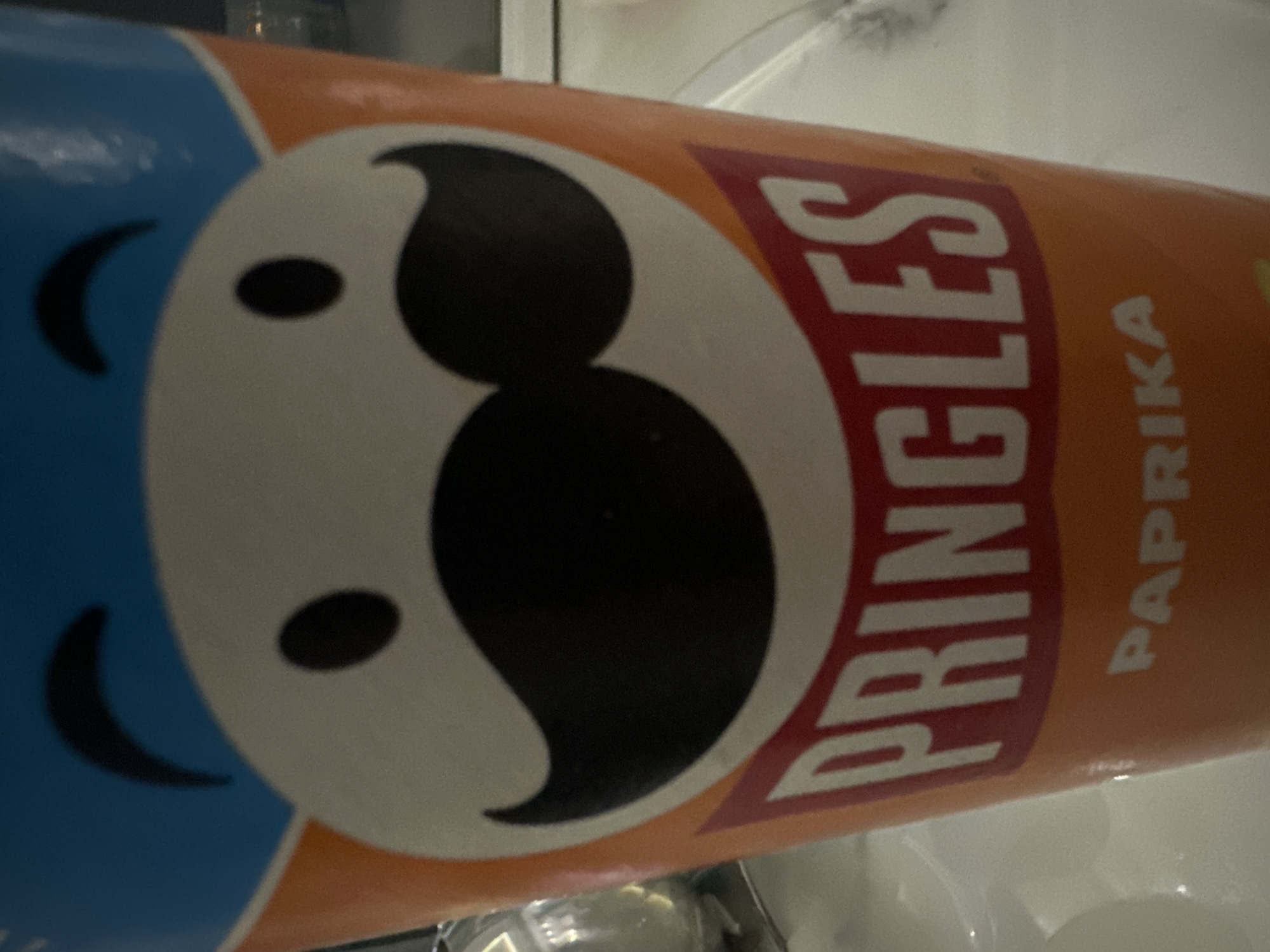 The image is a close-up shot of a Pringles can, specifically the "Paprika" flavor. The can is predominantly orange with a white section featuring the Pringles mascot's face, which includes black eyes and a black mustache. Above the white section is a blue band. The word "PRINGLES" is written in large white letters on a red background below the mascot's face, and "PAPRIKA" is written in smaller white letters on the orange part of the can. The lighting is somewhat dim, and the can is placed on a white surface that appears to be a table or countertop. The background is blurred, but suggests an indoor setting, possibly a kitchen or dining area. There are no people visible in the image. The image does not provide any information or context related to the city of Asenovgrad, Bulgaria.