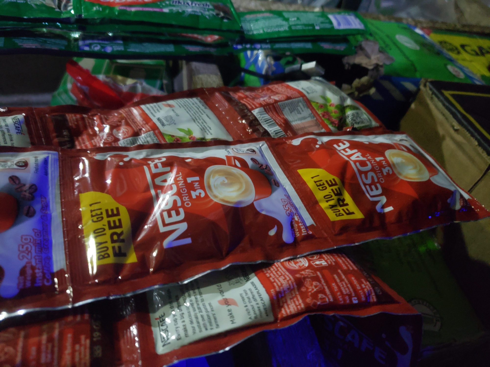 This is a close-up, eye-level, medium shot of several individually packaged Nescafe coffee sachets hanging vertically in what appears to be a stall or small shop. The sachets are predominantly red and white with prominent "Nescafe" branding. One sachet in the foreground displays a yellow sticker with the text "BUY 10, GET 1 FREE". The background is blurred and dark, with some green packaging visible on the right. The scene is indoors or under a sheltered area, with artificial lighting. The context of Jos, Nigeria, is not explicitly depicted in the image itself, but it is provided as the location. The image captures consumer products for sale, with an emphasis on a promotional offer. There are no people or active interactions visible.