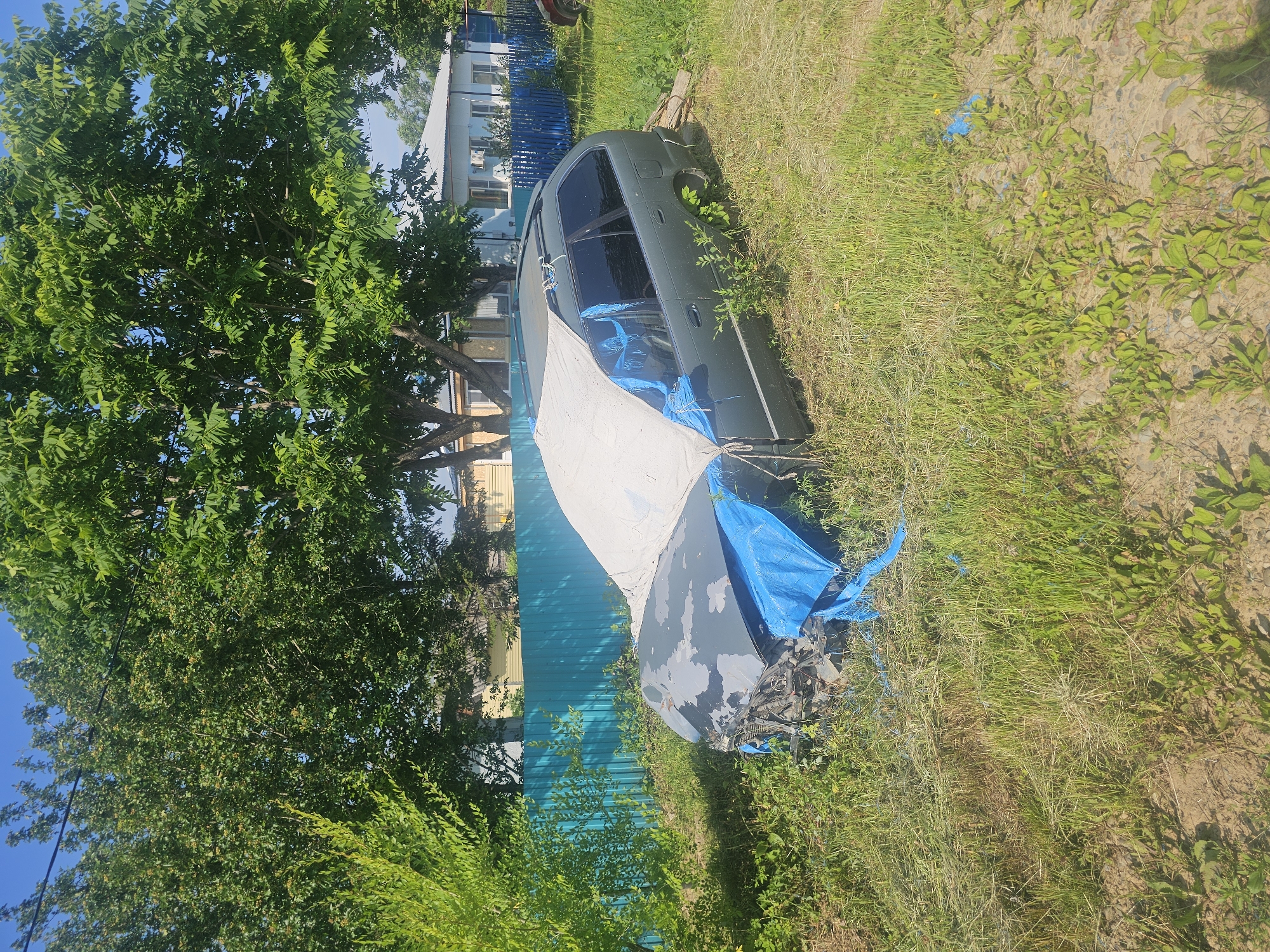 In Georgievka, Russia, a partially dismantled and covered dark green station wagon sits in a grassy, overgrown area. The front of the vehicle is exposed, showing significant damage and exposed engine components. Blue tarp material is visible covering parts of the front and sides, along with a larger light-colored tarp draped over the hood and windshield. The car is positioned near a bright turquoise metal fence, with residential buildings and trees visible in the background under a clear blue sky. The overall scene suggests the vehicle is abandoned or undergoing significant repair/disassembly outdoors.