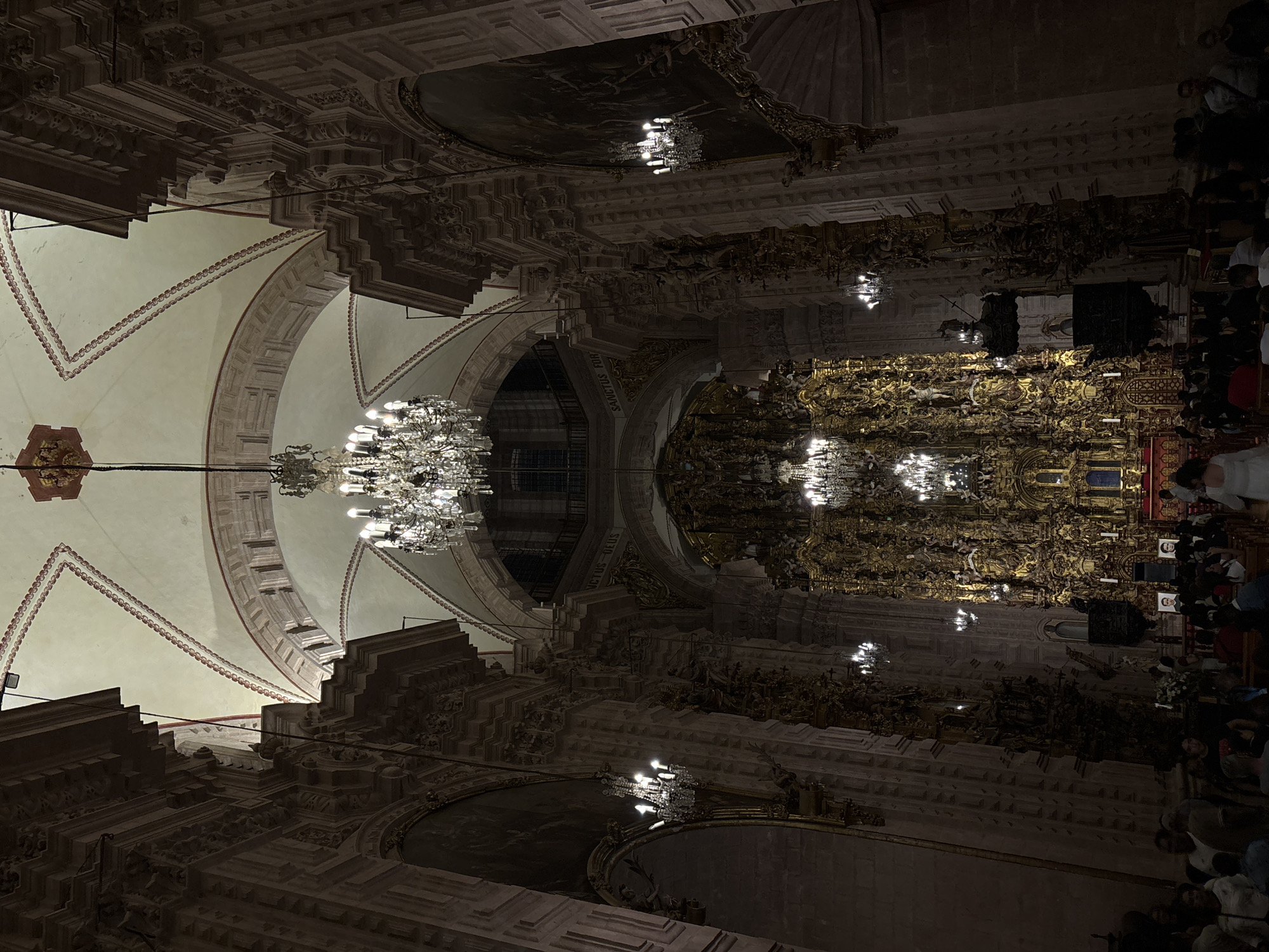 The image shows the interior of the Santa Prisca Church in Taxco, Mexico. The view is an upward angle, capturing the church's ceiling, arches, ornate gold altar, and chandeliers. The ceiling features detailed architectural elements. The altar is gold and intricately designed. People are present in the background.