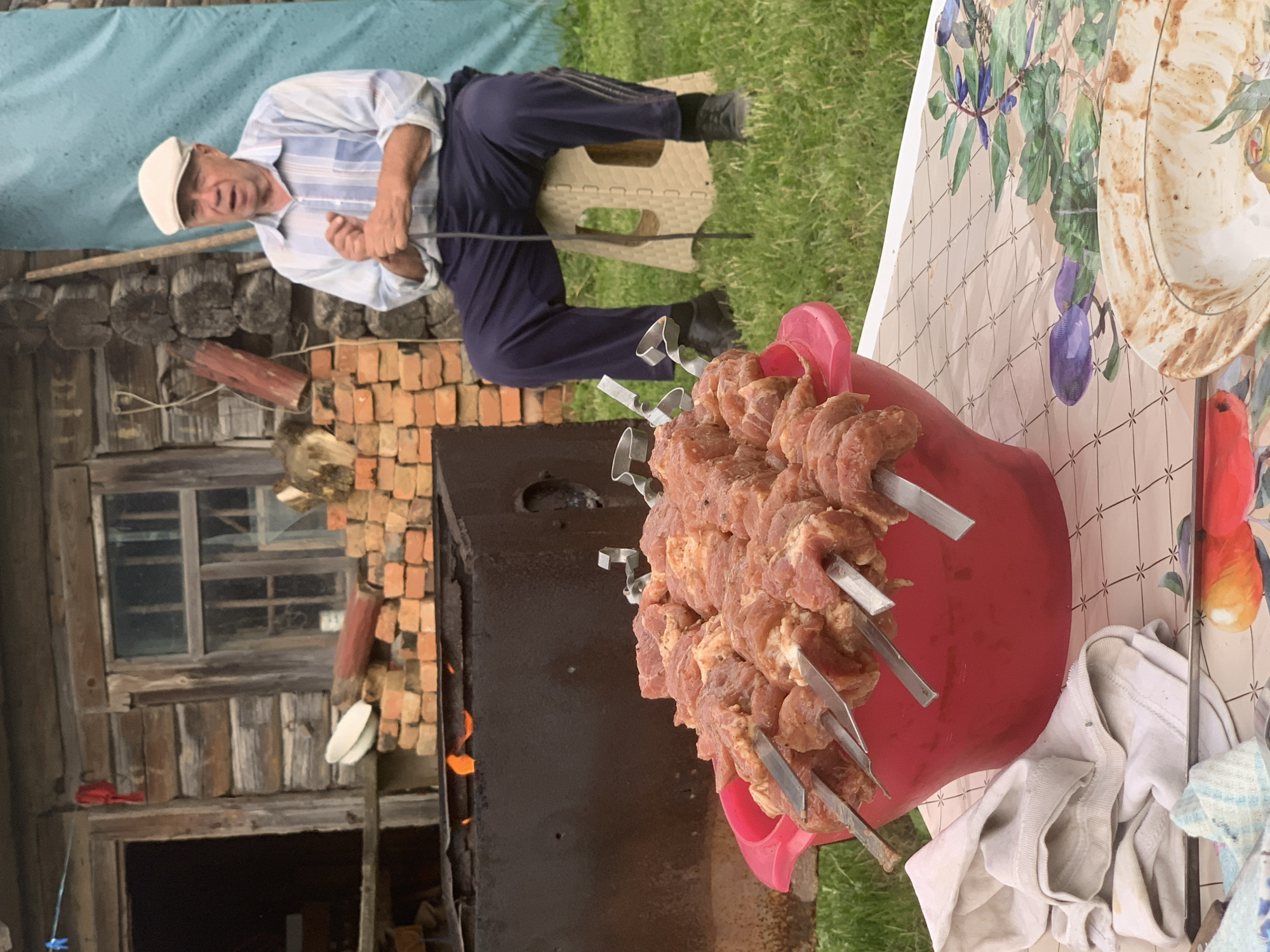 In Відзаўскі сельскі Савет, Belarus, an elderly man in a blue and white striped shirt and a white cap sits on a beige folding stool, holding a walking stick. In the foreground, a large red plastic bowl overflows with raw meat skewers, ready for grilling. Behind the skewers, a metal barbecue grill is visible, with flames flickering inside. The scene is set outdoors, with a rustic wooden building in the background, piles of bricks, and lush green grass. A patterned tablecloth covers a table in the foreground, with a dirty plate and some folded cloths on it.