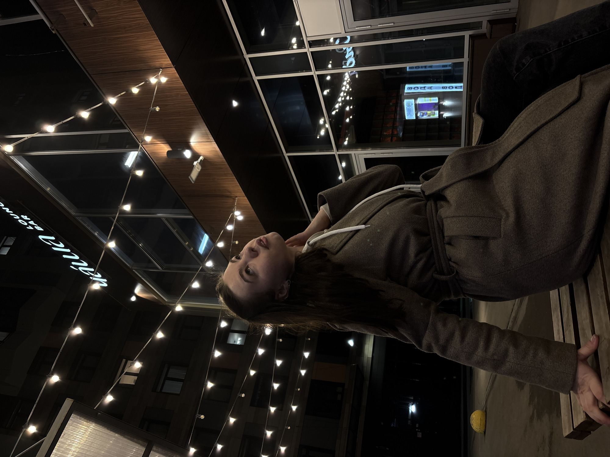 A young woman in a brown coat poses outdoors at night in Perm, Russia. The strings of lights and building signs are distinctive. The woman is on a bench. Her gaze is directed upward and toward the building.
