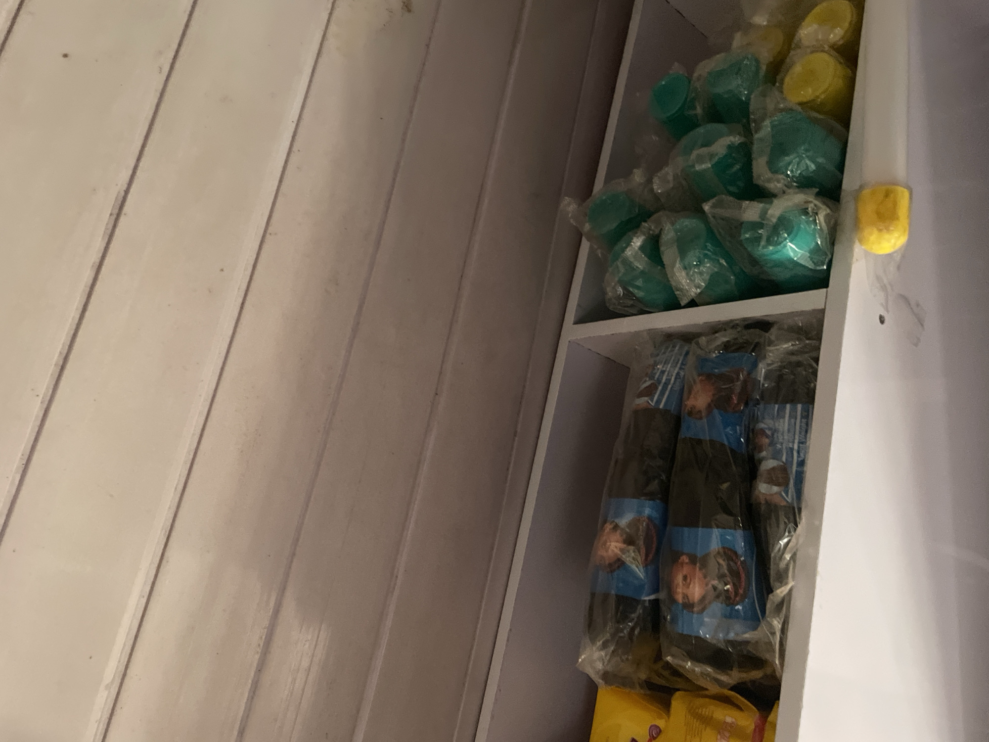 The image shows a shelf in what appears to be a retail or storage space, likely in the city of Durumi, Nigeria. The shelf is stocked with various items. On the left side, there are packages wrapped in clear plastic, with some featuring blue and black print that includes images of what seem to be food items. To the right of these, a section of the shelf contains a collection of teal-colored cups, also individually wrapped in plastic. Further to the right, several yellow items, possibly lids or small containers, are visible. A single yellow object, possibly a piece of fruit or a small toy, rests on the shelf below, near the front. The background is a wooden, slatted ceiling. The lighting is dim, and the overall scene is cluttered, suggesting a place where goods are stored or sold. There are no people visible in the image.