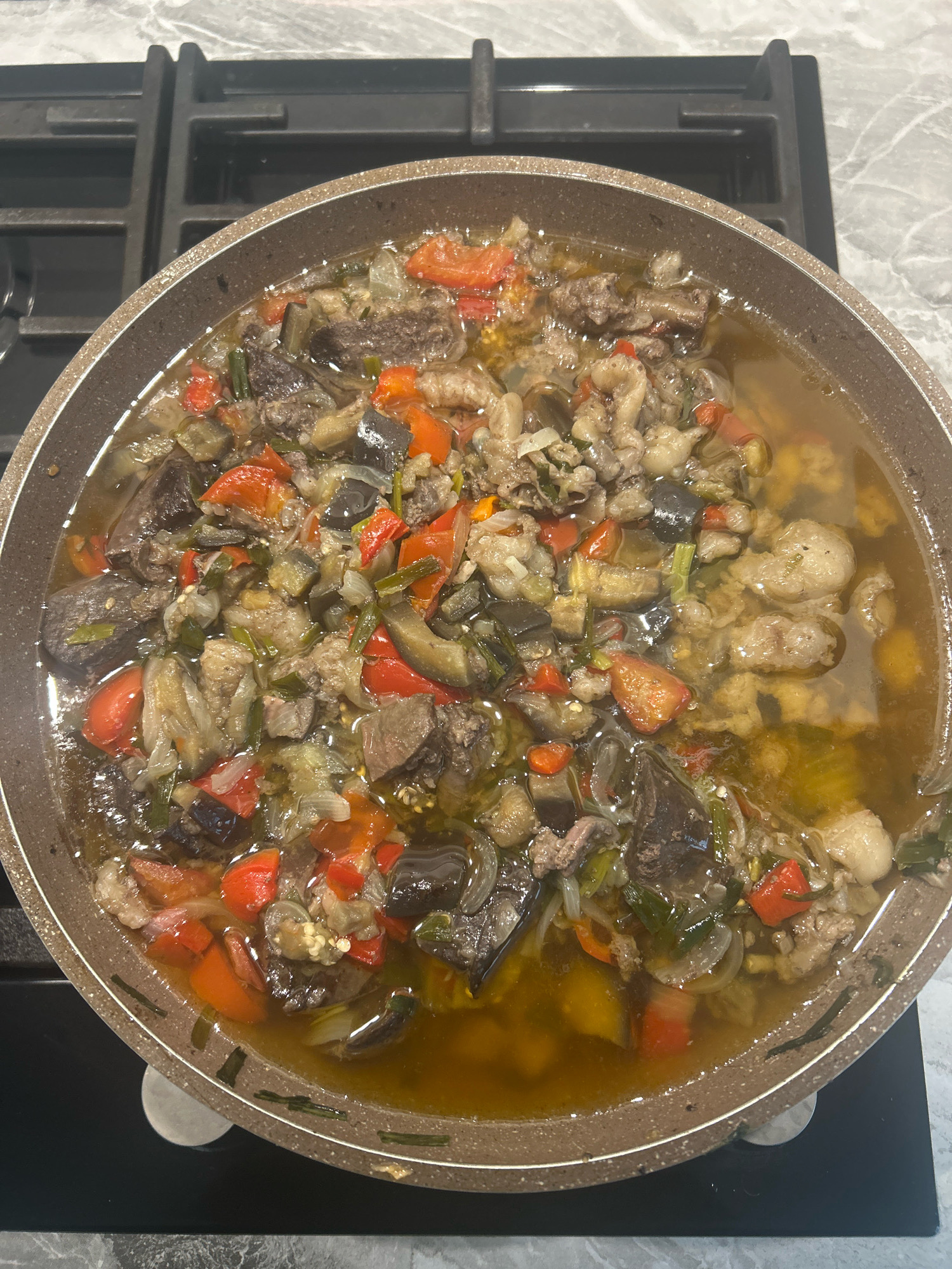 An overhead, close-up shot shows a brown-rimmed pan filled with a stew-like dish, likely a vegetable and meat ragout. The dish contains chunks of eggplant, red bell peppers, onions, and pieces of meat in a yellowish, slightly oily broth. The pan is placed on a black stovetop with a marble-patterned countertop in the background. The scene is an indoor setting, specifically a kitchen. There is no indication of the location being in Grozny, Russia.