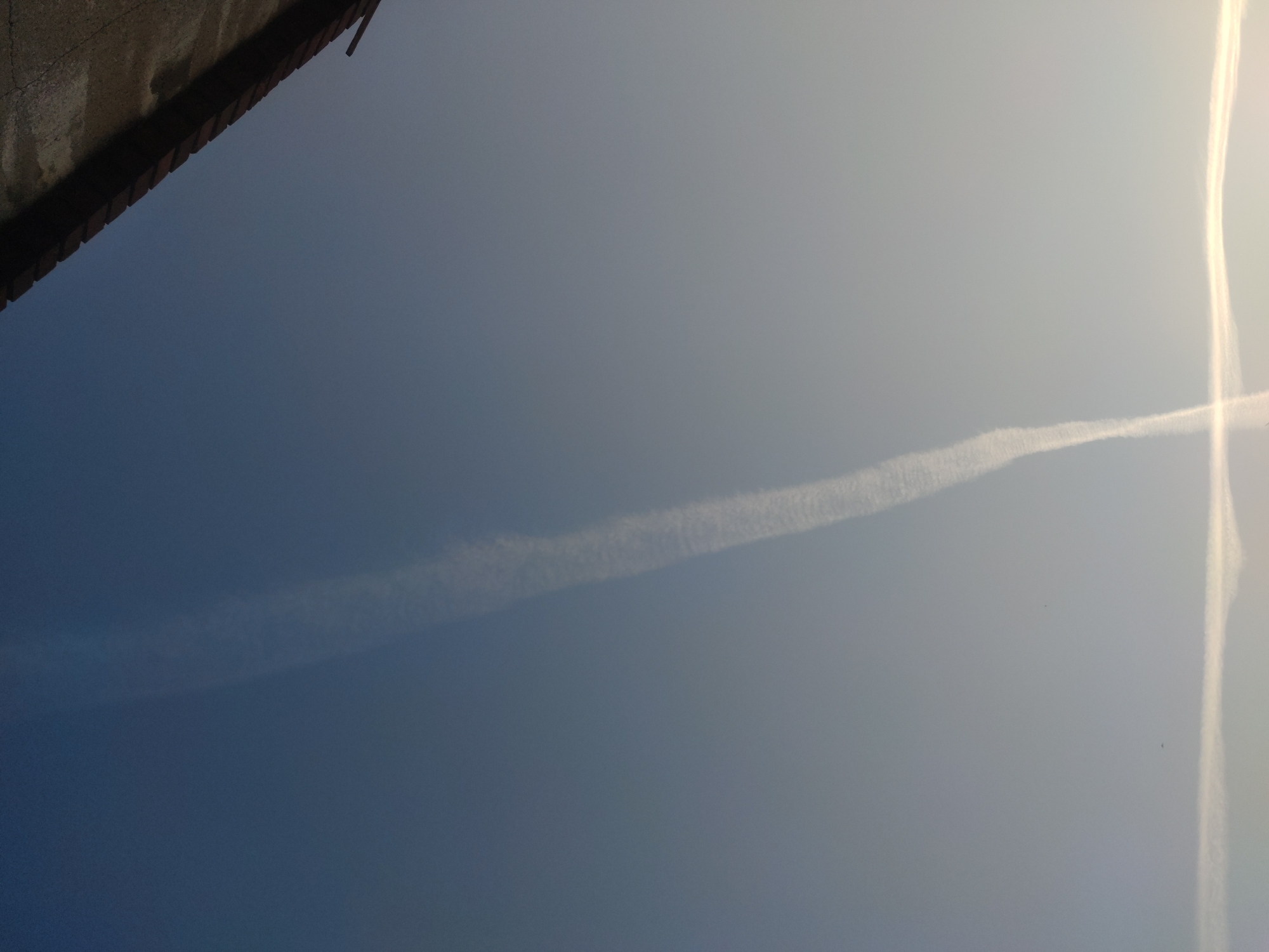 The image shows a view of the sky with contrails, one appearing as a horizontal line and another as a vertical line, possibly intersecting. A portion of a building with a bricked eave or edge is visible in the upper-left corner of the frame. The sky transitions from a darker blue on the left to a lighter, more washed-out tone on the right. The location cannot be determined as Barcelona based on the available visual data.