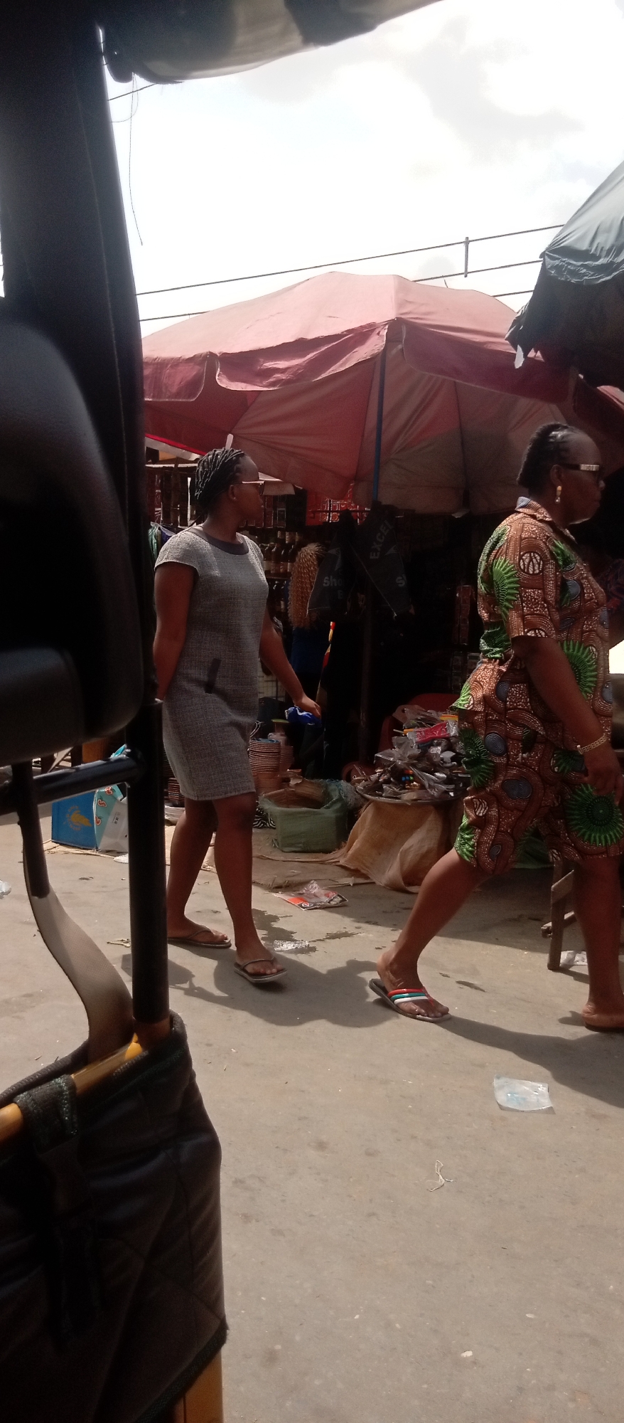 The image depicts a street scene in Onitsha, Nigeria. Two women are walking on the sidewalk. The woman on the left is wearing a gray dress and flip-flops. The woman on the right is wearing a brown and green patterned outfit and sandals. They are in front of a market stall covered by a red umbrella. Various items are displayed for sale, including shoes and other goods. The view is partially obstructed by a black vehicle part. The weather appears sunny. The ground is concrete. There is litter on the ground.