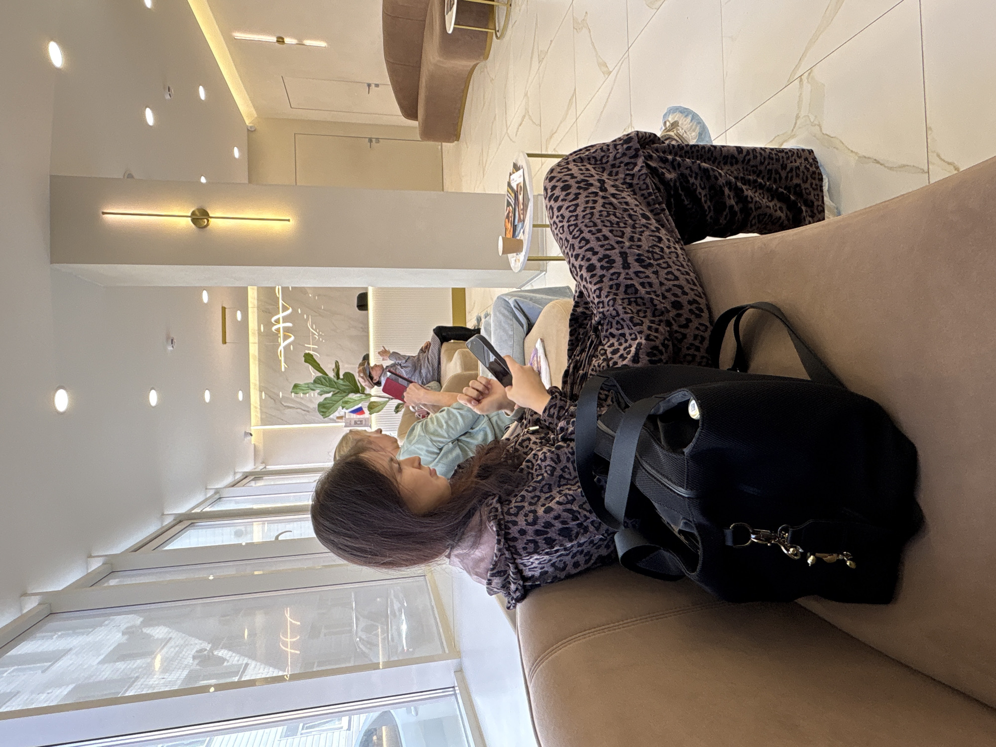 The scene depicts the interior of what appears to be a waiting area or lounge. A woman with long dark hair, wearing a leopard-print patterned outfit, is seated on a light brown upholstered sofa. She is holding a smartphone, possibly looking at the screen. A large black bag sits next to her on the sofa. In the background, other people are seated and appear to be using their own phones. Natural light comes through a large window on the left side of the frame. The ceiling has modern lighting fixtures. The floor is light-colored with a marble-like pattern. The location is Perm, Russia.