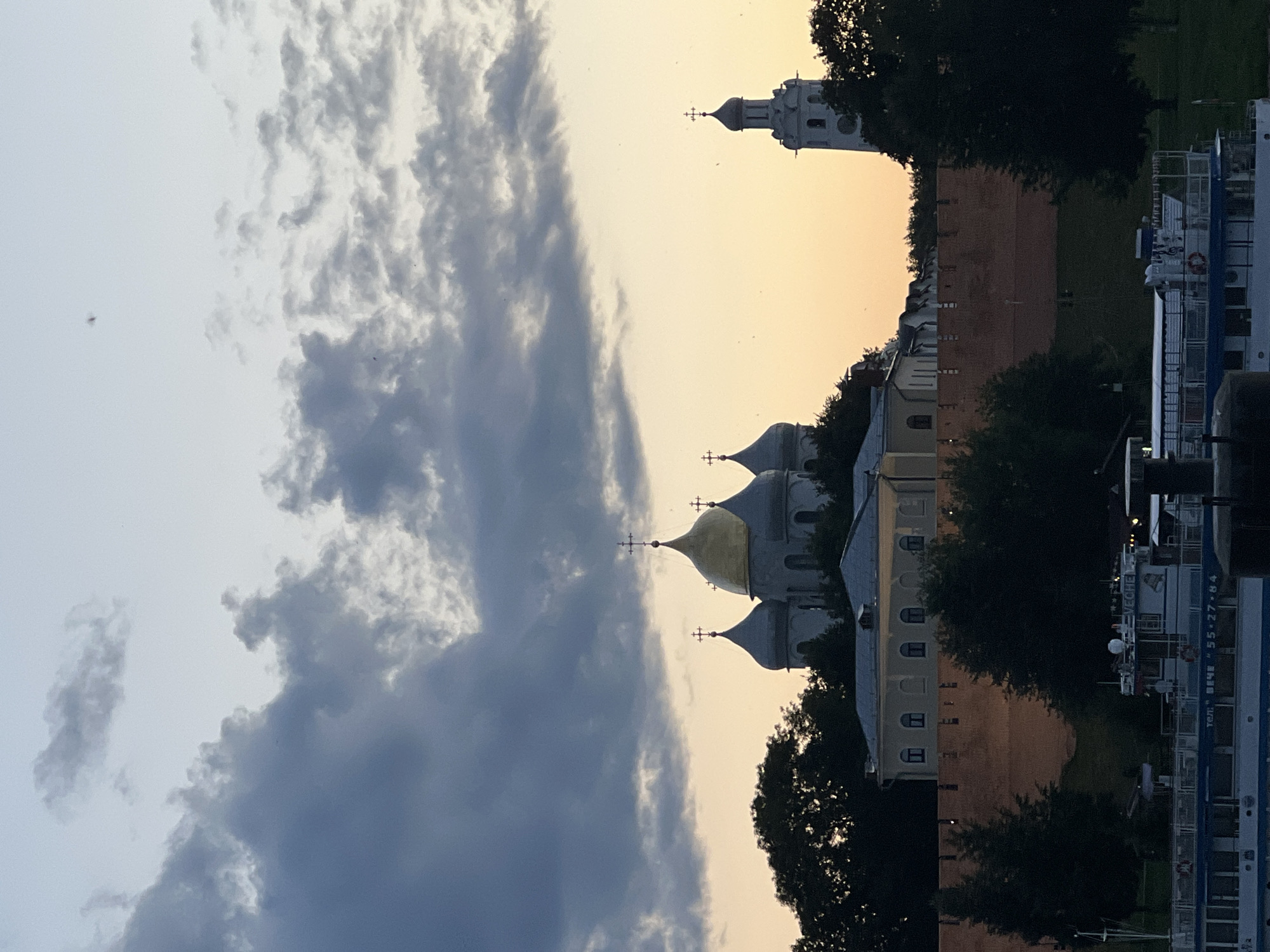 This image depicts a view of the Veliky Novgorod Kremlin, also known as the Detinets, a historic fortified complex in Veliky Novgorod, Russia, as dusk approaches. The main architectural feature is the Saint Sophia Cathedral, recognizable by its multiple domes, one of which is golden. Adjacent to the cathedral, a tall bell tower with a white spire stands silhouetted against the pale orange and grey sky. A red brick wall, characteristic of the Kremlin's fortifications, forms a prominent horizontal line across the mid-ground, with dark green trees partially obscuring it. In the foreground, a white boat with the text "VECHE" is visible on the water, suggesting the view is from a river. The sky is filled with dramatic clouds, ranging from dark grey to lighter shades, interspersed with hints of the setting sun's warm light. Several small, dark specks, possibly birds or insects, are scattered across the sky.
