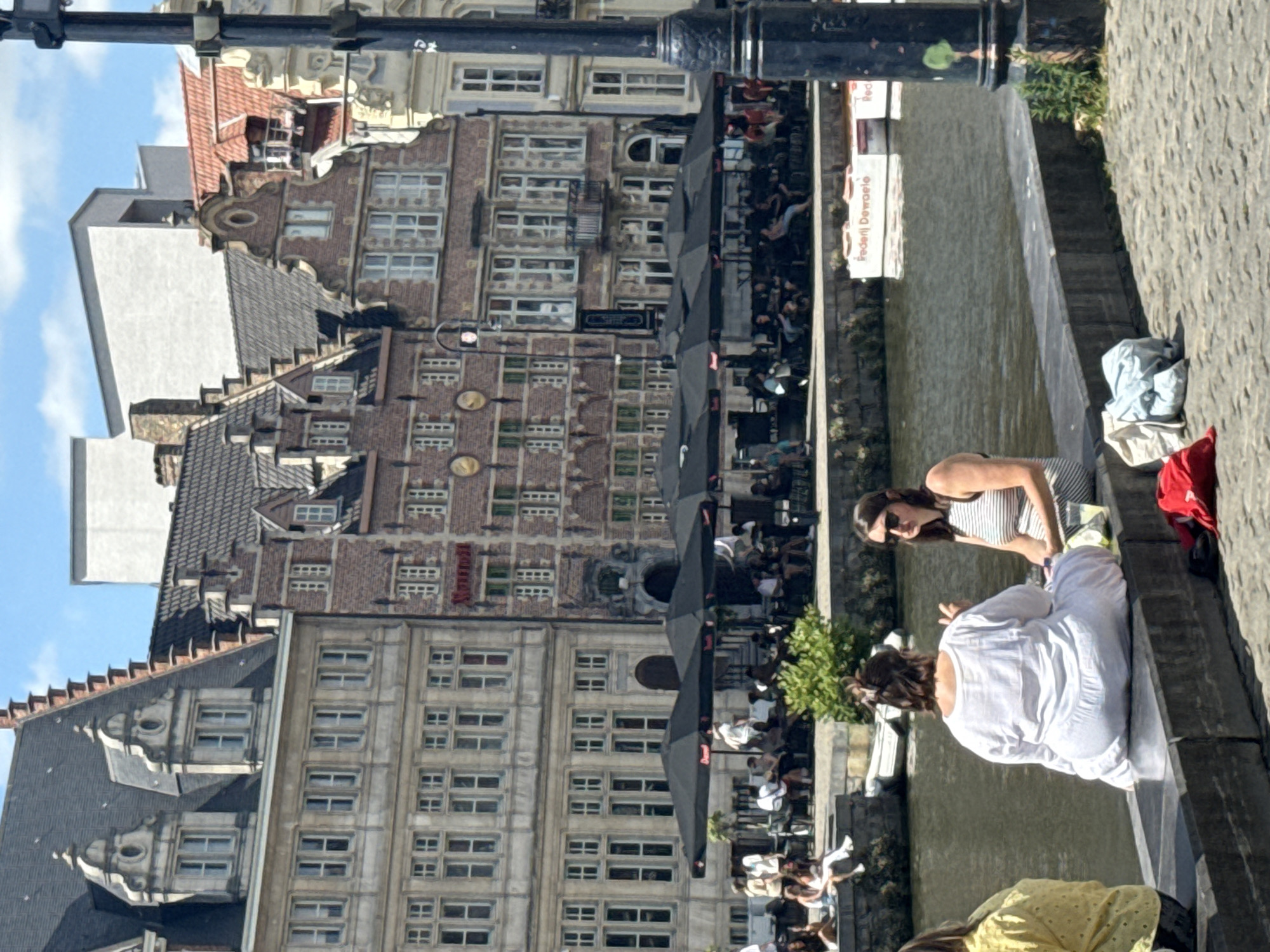 In this media file, people are enjoying a sunny day by a canal in Ghent, Belgium. The scene depicts a charming canal-side with historic brick buildings adorned with numerous windows. Several individuals are seated on a stone ledge by the water, with some appearing relaxed and others engaged in conversation.
One woman, wearing a striped top and sunglasses, is seated, while another, in a white long-sleeved shirt, is in the foreground, facing away from the camera. A person in a yellow top with a polka dot pattern is also visible. Clothing items, including a blue shirt and a red jacket, are casually placed on the ledge.
The activity appears to be a leisurely outing, possibly a break from sightseeing or a casual get-together. The presence of umbrellas over an outdoor seating area suggests a cafe or restaurant. The lighting indicates it is daytime, likely with pleasant weather given the outdoor activities and attire. The architecture of the buildings, with their distinctive gabled roofs and brick facades, strongly points to a European city, consistent with the provided location context of Ghent, Belgium.
A sign with text is partially visible on a building, reading "Frederi Dewaele" and "Red." The overall mood conveyed is one of relaxation and enjoyment of a beautiful day in a historic urban setting.