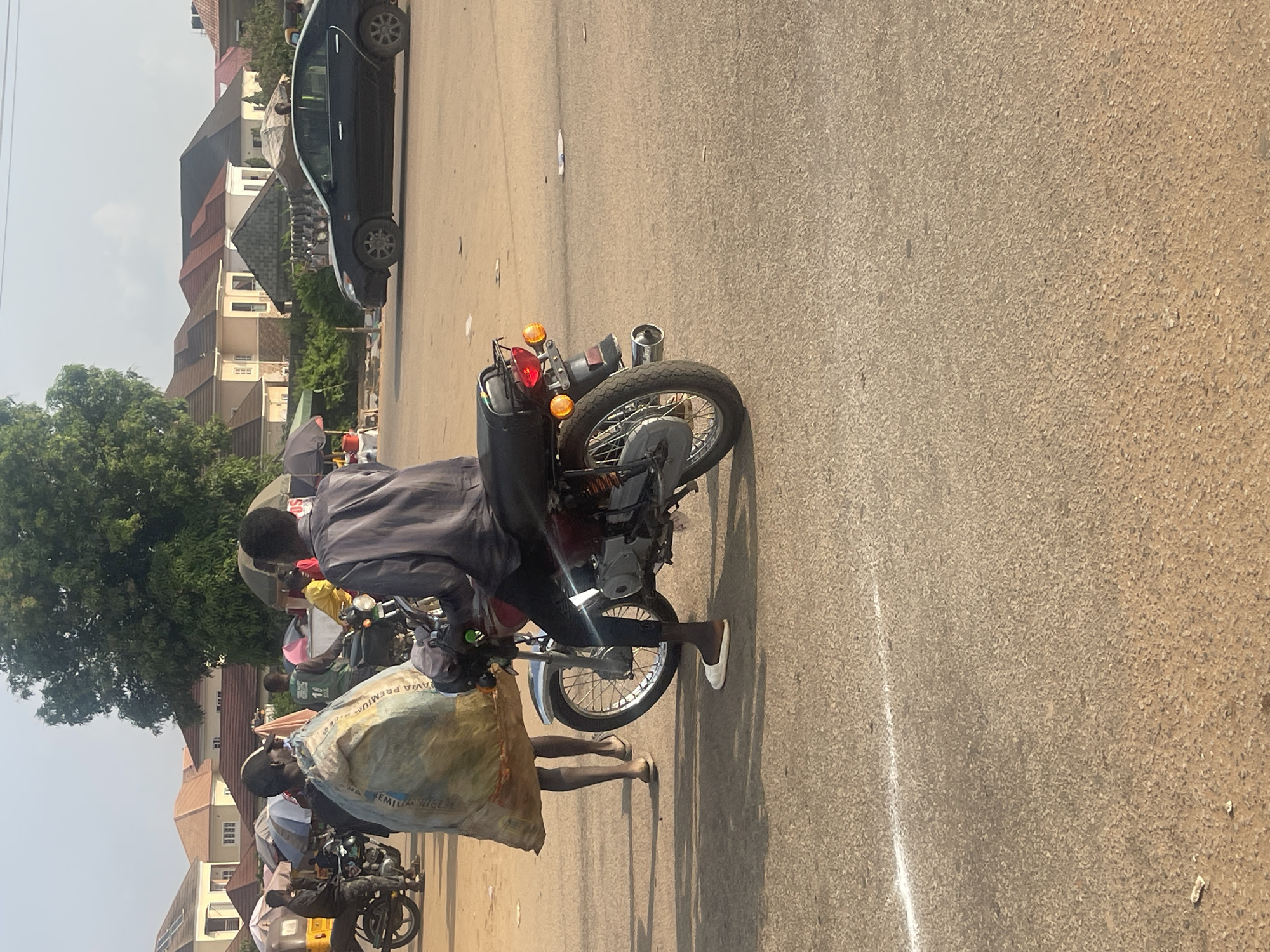 In Galadimawa Village, Nigeria, a street scene unfolds with a focus on a motorcycle and its occupants. A man, dressed in a gray shirt and dark pants, is seated on the motorcycle, which is positioned on a paved road. He is wearing white shoes. Another individual, also wearing dark clothing and a cap, is seen carrying a large, light-colored sack. This sack appears to be made of woven material and has red and blue lettering on it, with the visible text reading "AVIA PREMIUM RICE". The motorcycle itself has a red tail light and orange turn signals, and appears to be loaded with cargo, possibly within a black container behind the seat.
In the background, several houses with distinctive architectural styles, featuring peaked roofs, are visible. Green trees dot the landscape, suggesting a warm climate. The road is asphalt, marked with a white line on the right side. The lighting suggests it is daytime, with clear skies. The overall impression is one of everyday activity and commerce in a Nigerian village setting.