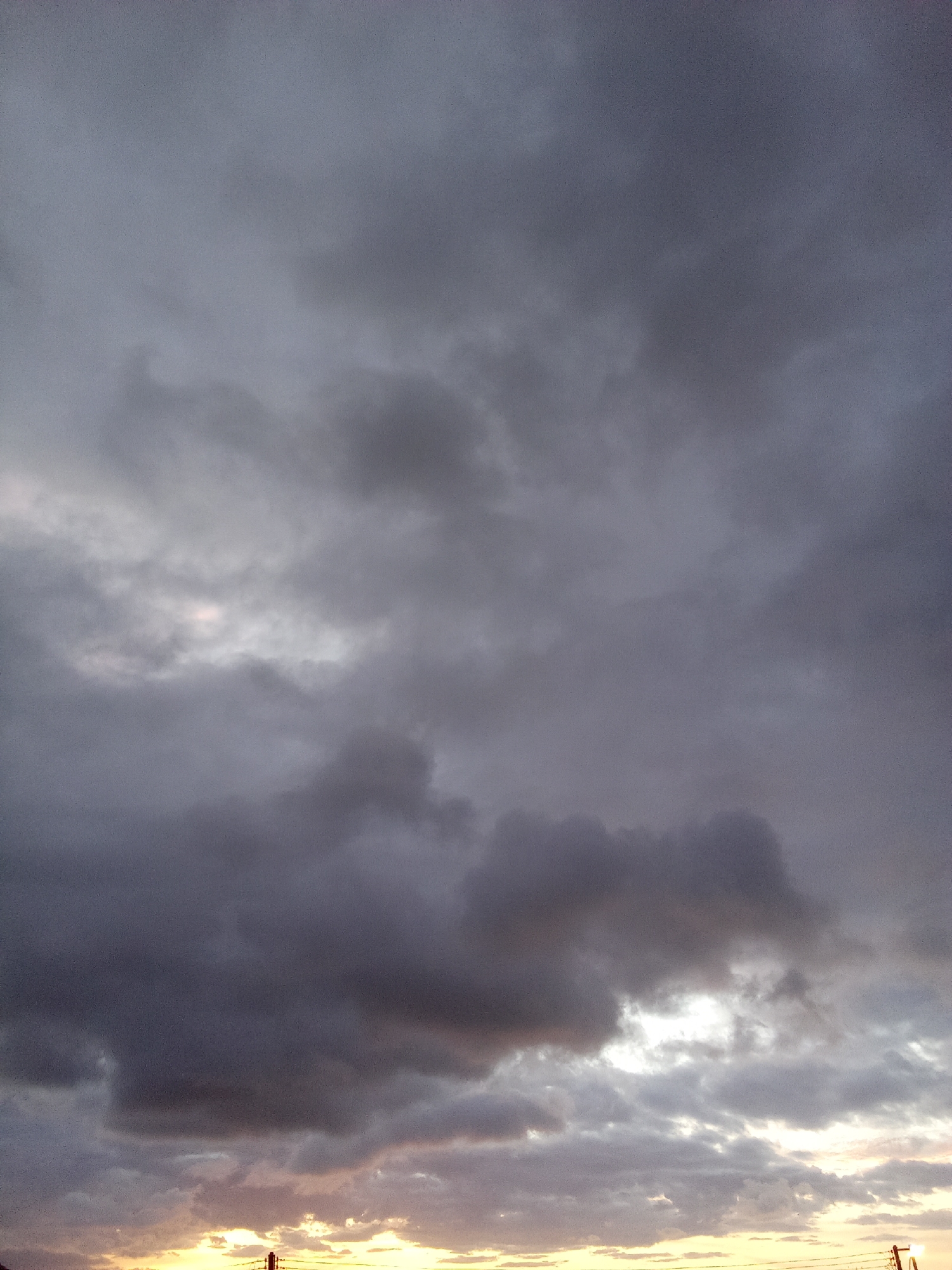 A low-angle, vertical, outdoor shot depicts an overcast sky at sunset. The sky is predominantly filled with dark, heavy clouds, varying in shades of gray, obscuring most of the view. At the bottom of the frame, a faint orange and yellow glow of the setting sun peeks through the cloud cover, suggesting it is near the horizon. The horizon is undefined, as the cloud cover is low, with some utility poles silhouetted against the bright light. The location is identified as the city of Juan Griego, Venezuela, although there are no specific landmarks to confirm the precise location. There are no people or subjects immediately visible. The focus of the scene is the dramatic interplay of light and shadow in the cloud formation during the late day in Venezuela.