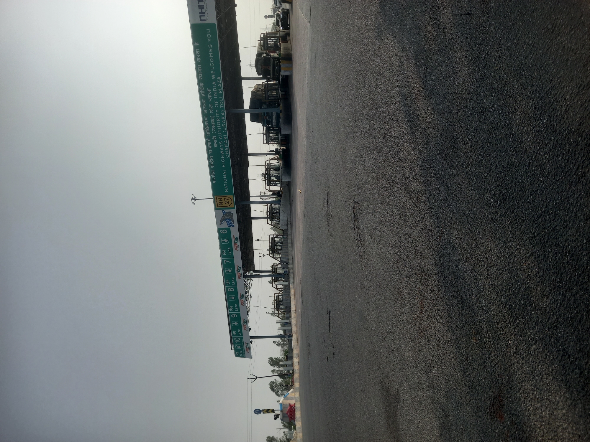 The image depicts a toll plaza in Usargaon, India, specifically the Chamari (Usaka) Toll Plaza on National Highway 27. The plaza is marked by overhead signs indicating lane numbers (6 through 10) and accepting FASTag payments. The text on the sign states "National Highways Authority of India Welcomes You." Several vehicles are present under the tollbooths. The foreground shows a paved road surface. The sky is overcast.