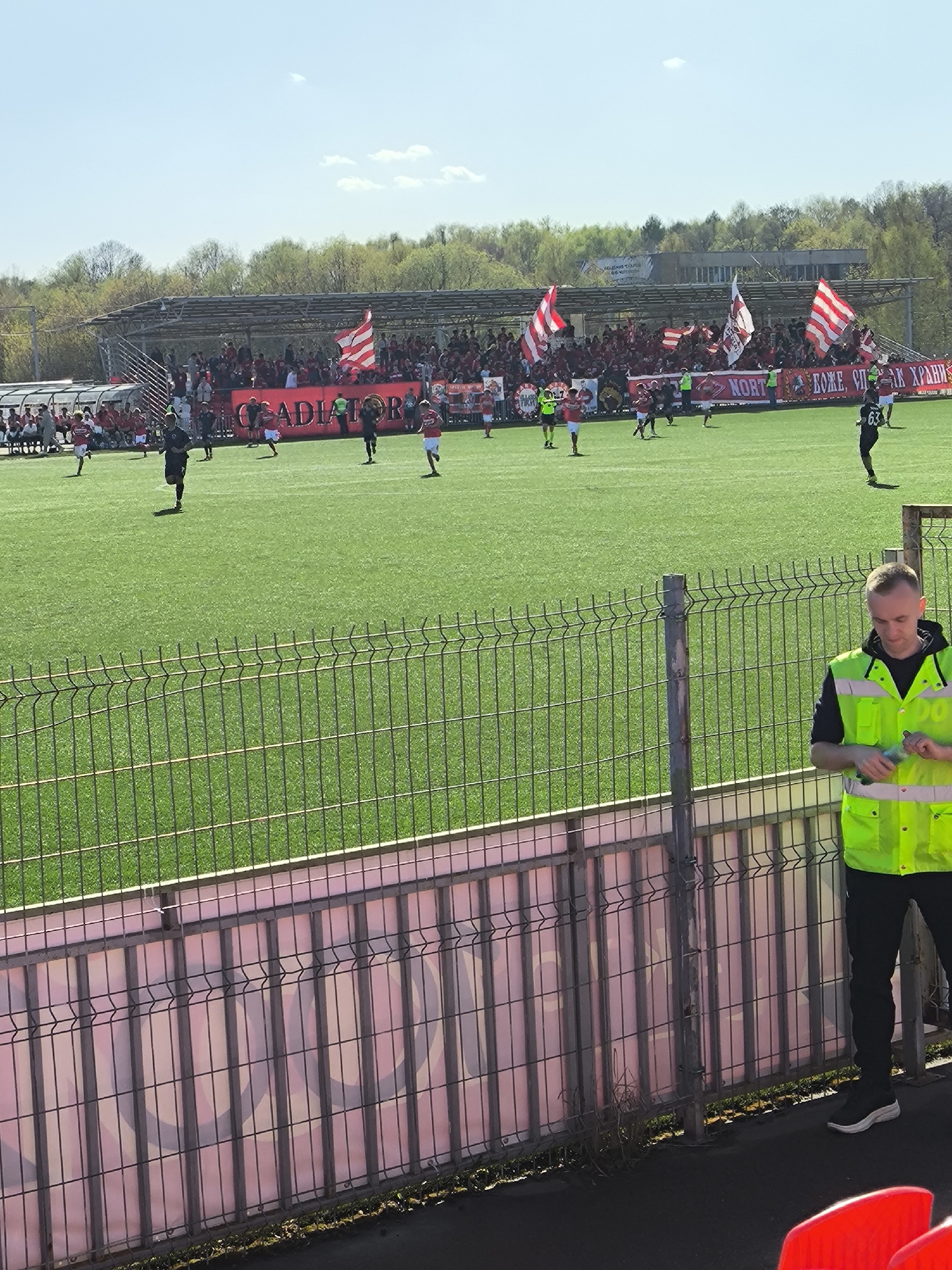 The image depicts a soccer match in progress at Spartakovets Stadium in Moscow, Russia, under a clear, bright sky. The green playing field is populated by players engaged in a game. Spectators fill the bleachers, waving flags and banners bearing the word "GLADIATOR," supporting their team. A metal fence separates the field from the viewing area. In the foreground, a security guard wearing a neon yellow vest stands near the fence.