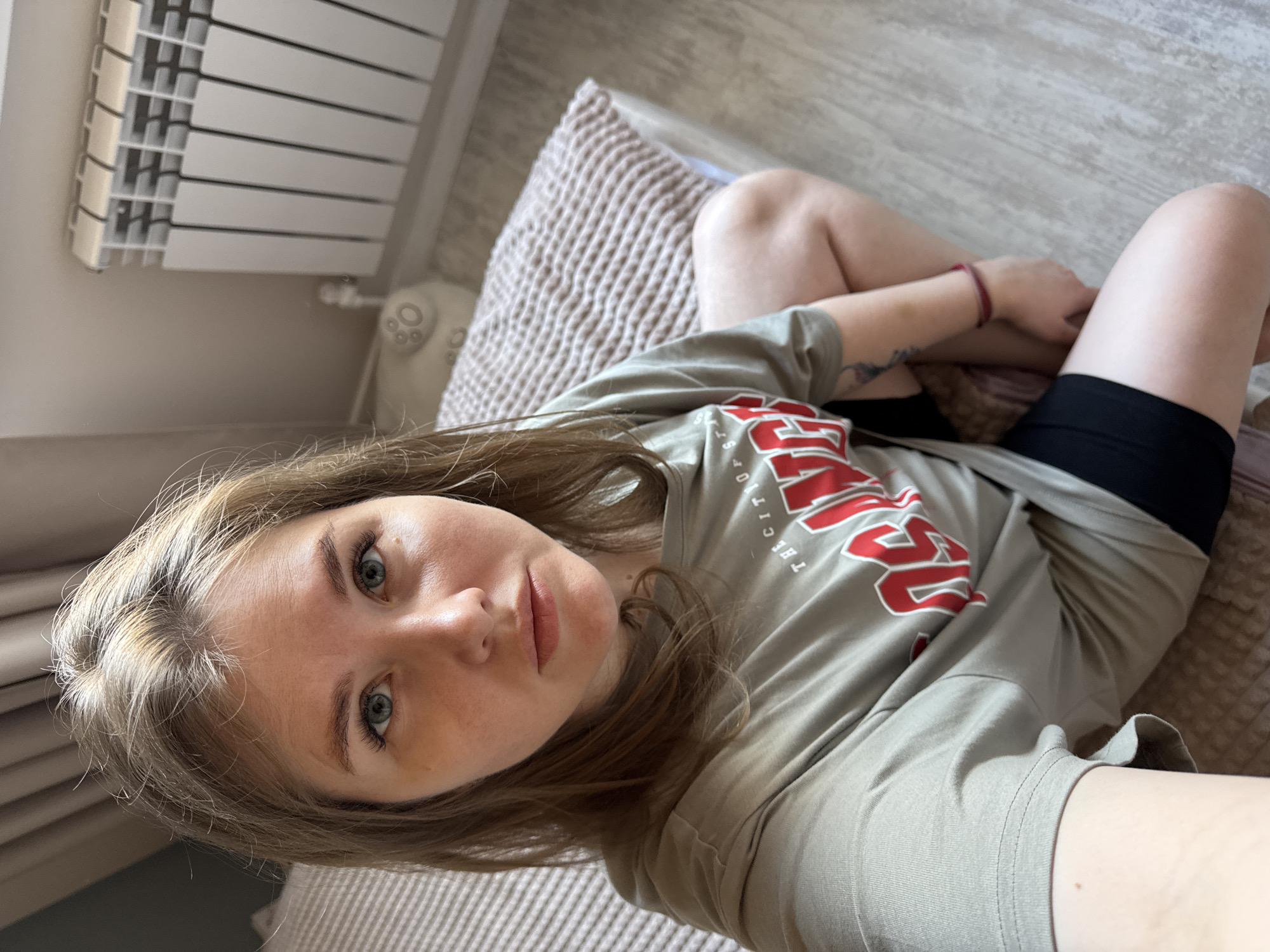A young woman with fair skin and shoulder-length light brown hair is lying on a bed with light brown quilted bedding. She is wearing a light green t-shirt with red and white writing and black shorts. She is looking directly at the camera with a slight smile. Her legs are crossed at the knees. A white radiator is visible in the background near a light gray wall. The floor is light gray wood. The scene appears to be indoors, possibly a bedroom. The location in Gornyi (Dvurechenskoe S/P), Russia, is not definitively discernible from the image.