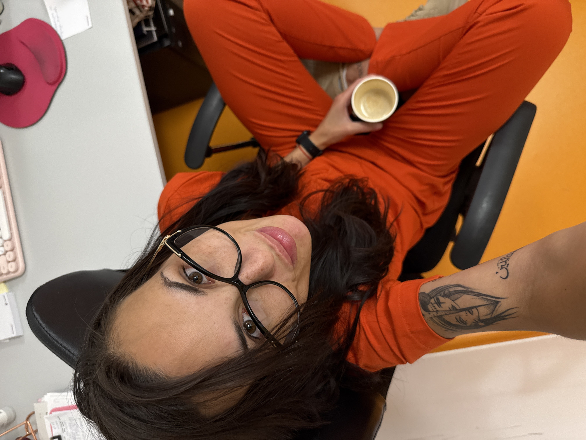 A woman with dark hair and glasses is sitting in a chair, wearing an orange jumpsuit. She is holding a cup. A tattoo is visible on her left arm. The location is not Pleven, Bulgaria. The environment appears to be an office, with a desk and computer peripherals visible.