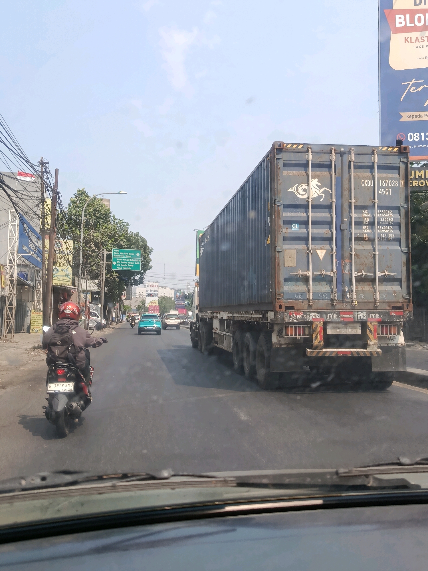 The image depicts a street scene in Bekasi, Indonesia, under a bright, clear sky. In the foreground, a portion of a vehicle's windshield and dashboard is visible. The main focus of the image is a large blue shipping container truck, prominently positioned in the right lane of the road. The truck's rear is visible, showing the container's markings, including "CXDU 167028 7 45G1," and weight specifications. The truck has multiple axles and appears to be moving.
In the left lane, a person on a black motorcycle, wearing a grey jacket, black pants, and a red helmet, is riding ahead of the truck. The motorcycle's license plate reads "B 3919 KMU." Several other vehicles are present on the road, including a light blue car in the middle lane and a white car further ahead.
The environment includes sidewalks with various buildings and businesses, some with signage in Indonesian. Trees and utility poles with a dense network of wires are also visible along the sides of the road. A green traffic signpost with directions to "Kranji," "Kantor H.K.d. Bekasi," "Summarecon Bekasi," "Tol Barat," and "RS. Hosana Insan Prima" is also present. A large billboard with text in Indonesian, including "BLO," "KLASTIK," and a phone number "0813," is located in the background. The overall impression is a busy urban street scene with moderate traffic.