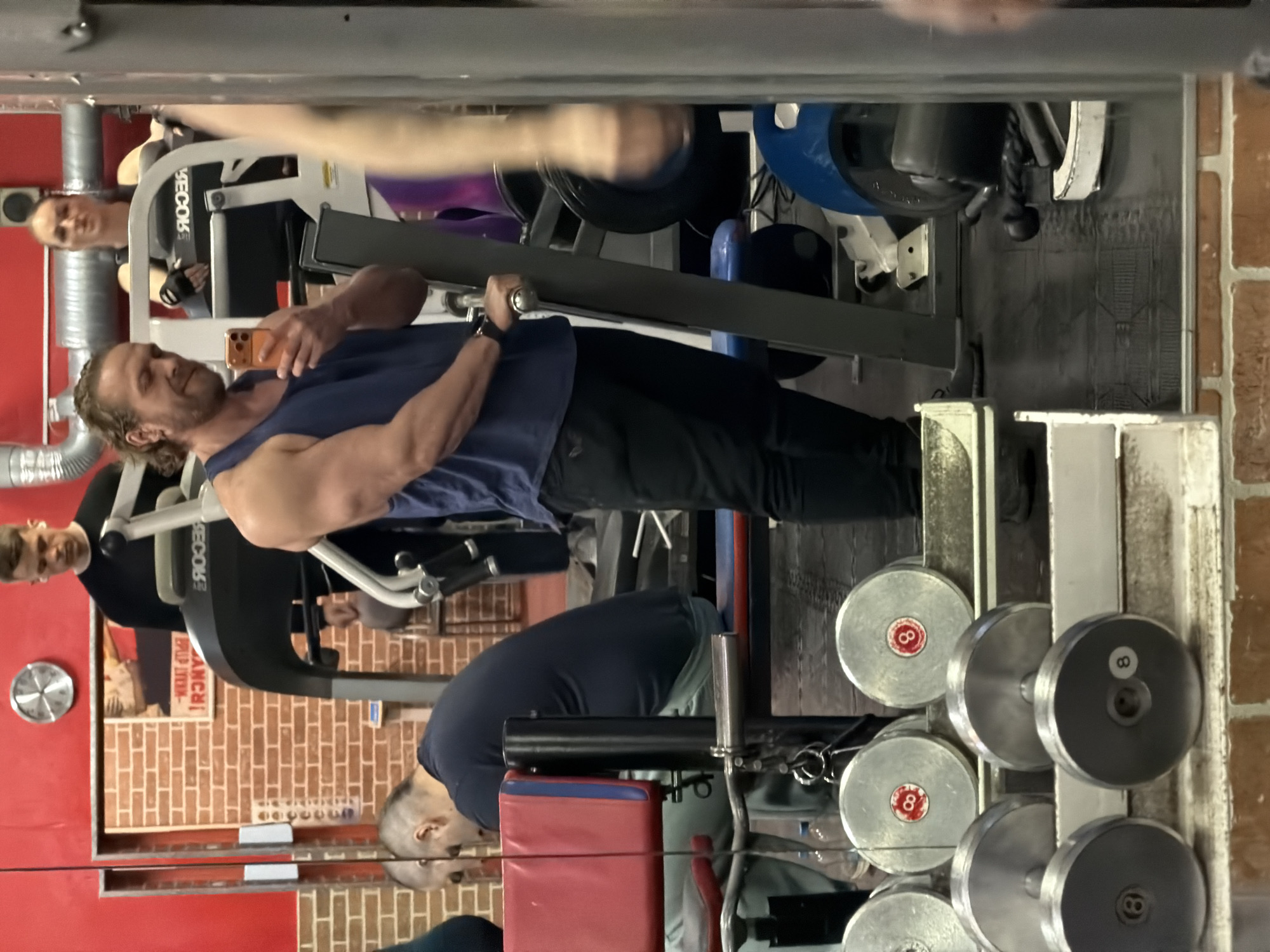 A mirrored view in a gym is observed. A man with long hair, wearing a blue tank top and black pants, is using a weight machine, while holding a phone in one hand. Another person is seated on the weight machine as well, and another person is standing behind the machine. Another person is seen bending over in front of the mirrors. Dumbbells are arranged on a rack. The setting appears to be a gym with red brick walls. The exact location in Saint Petersburg, Russia cannot be determined from this image alone.