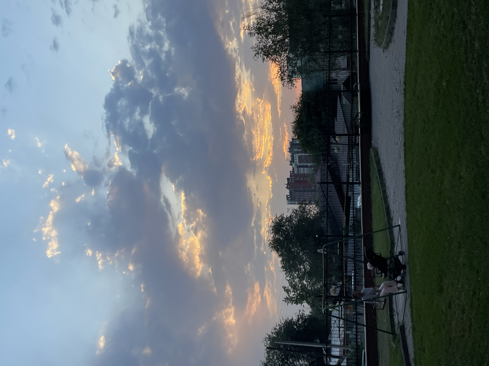 The image depicts a scene at dusk in Almaty, Kazakhstan, likely a residential area. The sky is filled with dramatic clouds, with patches of orange and yellow light suggesting the setting sun breaking through. In the foreground, a grassy area transitions to a paved courtyard with a playground. A woman is seated on a swing set, and a baby stroller is next to it. Behind the playground, trees and a fence line the perimeter. In the distance, apartment buildings are visible against the twilight sky, with the tallest building having a distinctive facade. The overall impression is a quiet evening in an urban environment.