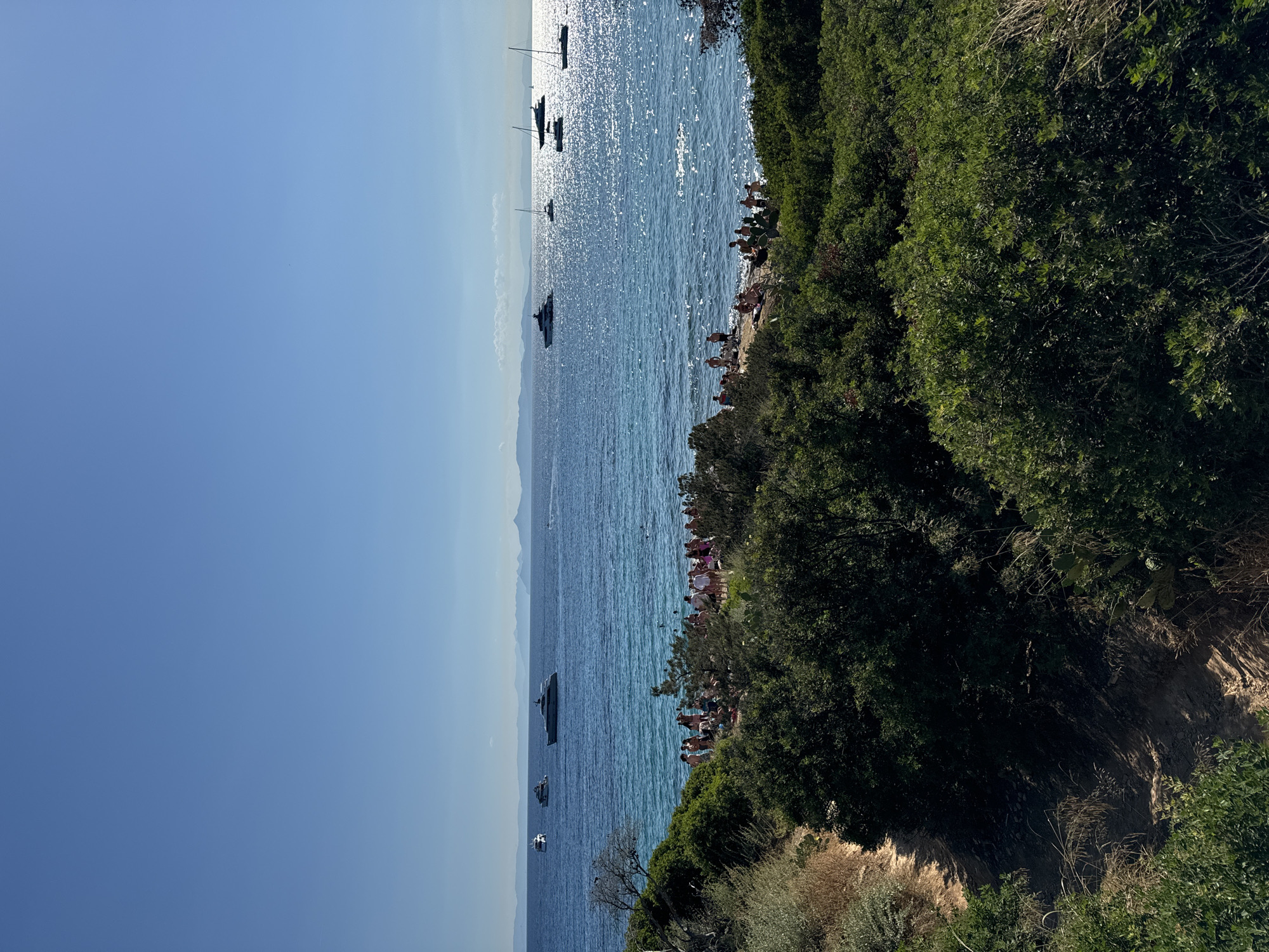 A scenic view of a coastal area in Vouliagmeni Municipal Unit, Greece, captures a sunny day at the beach. The foreground is dominated by lush green vegetation, with a dirt path winding through the foliage. Beyond the greenery, a crowded beach with numerous people enjoying the sun and sea is visible. The turquoise waters of the sea are dotted with several boats and yachts, and distant mountains form the horizon under a clear blue sky. The sun's reflection creates a shimmering effect on the water's surface.