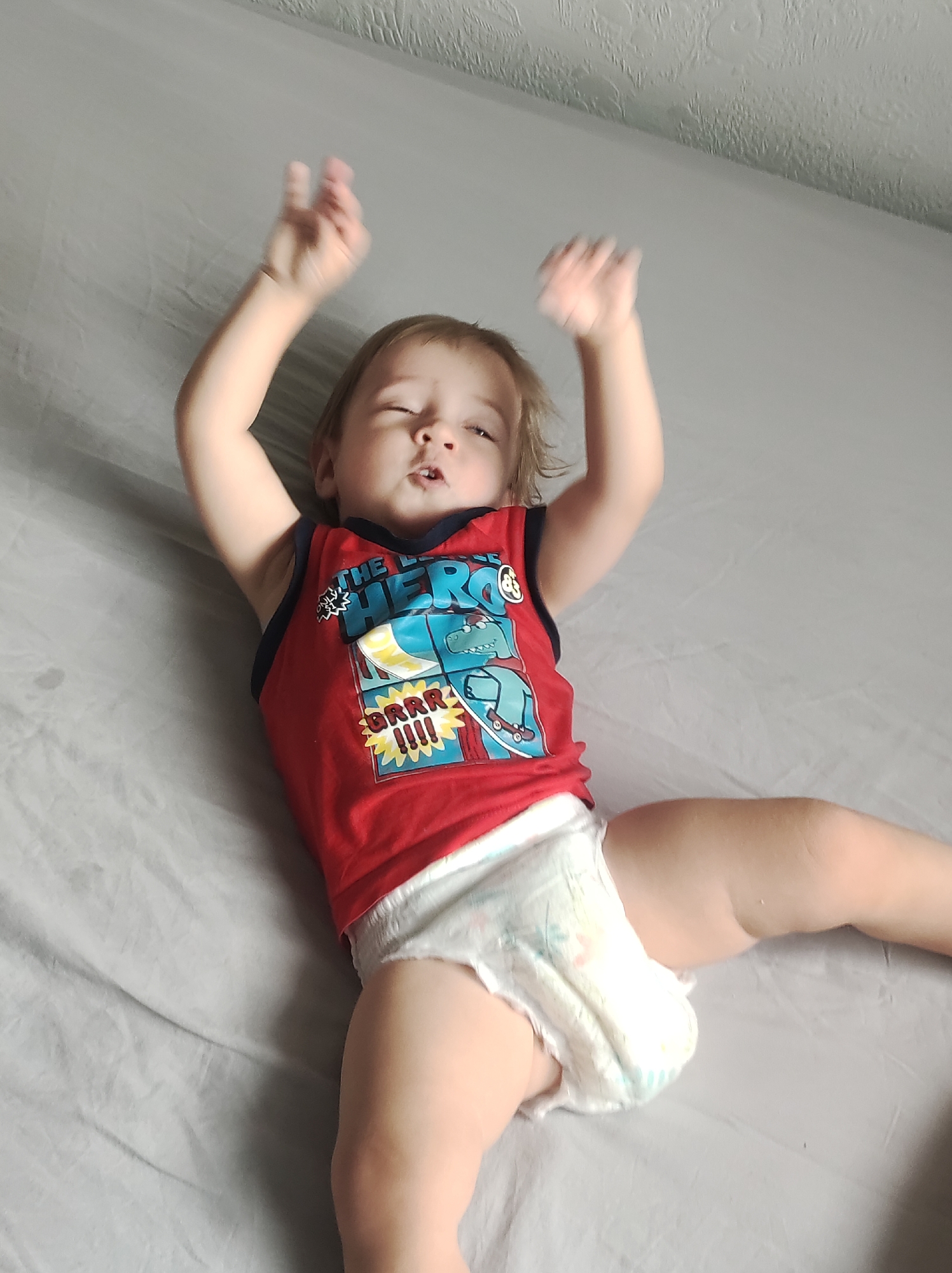 A toddler wearing a red tank top with a dinosaur graphic and a diaper lies on their back on a gray bedsheet. Their arms are raised. The photo was taken indoors. The location is unknown, but requested location of Chaikovsky, Russia could not be confirmed.
