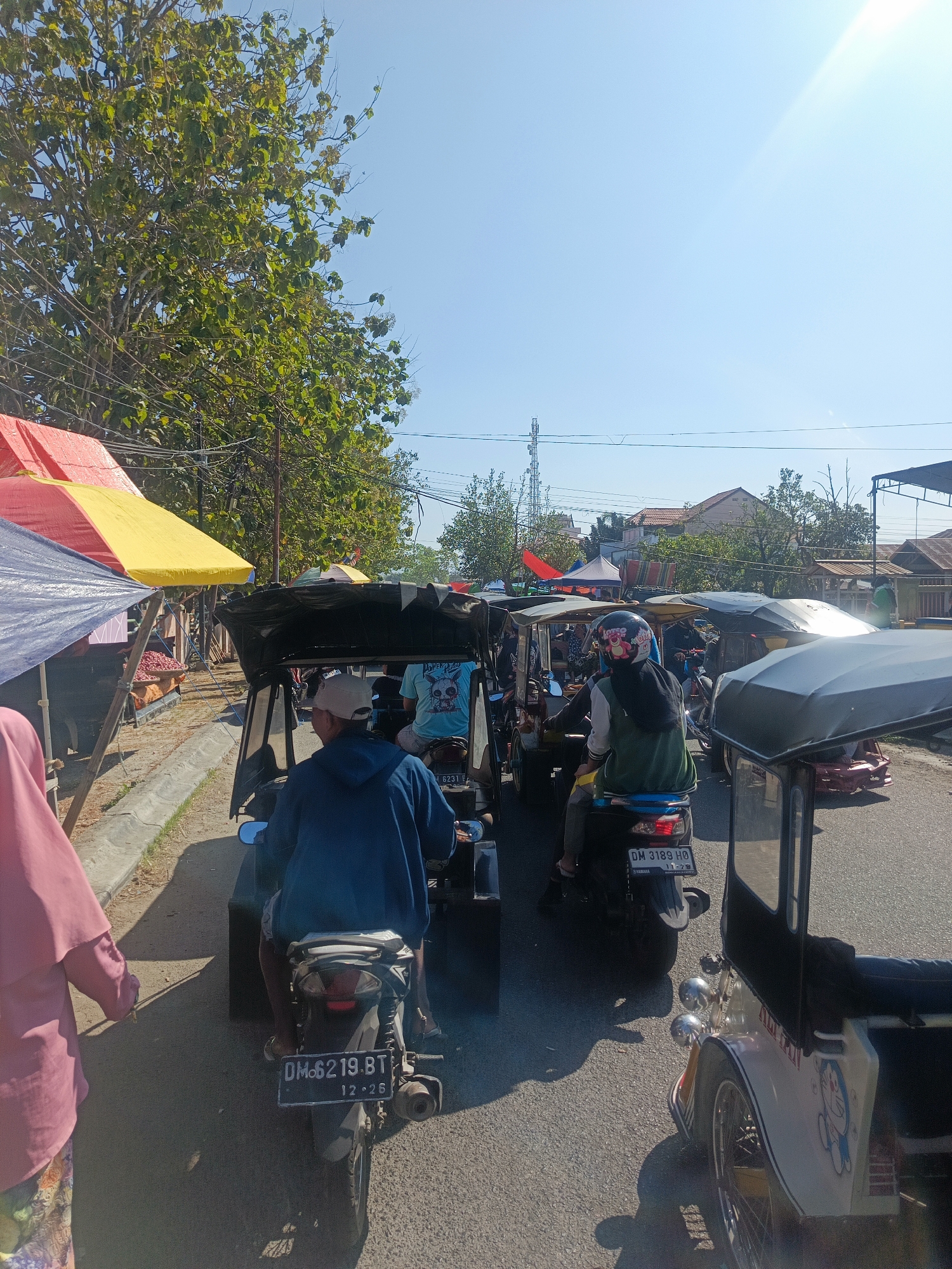 The image depicts a street scene in an outdoor market, likely in a tropical or sub-tropical region. The foreground is dominated by several three-wheeled vehicles, commonly known as "becaks" or auto-rickshaws, and motorcycles. One becak on the right has a white body with a cartoon character illustration. In the center, a becak with a black canopy is visible, with two individuals inside. Another becak, in the middle ground, is occupied by a person wearing a helmet. To the left, a motorcycle with license plate "DM6219.BT" is seen. A person wearing a pink headscarf and a colorful patterned skirt is partially visible in the lower left corner, walking away from the camera.
The background reveals a vibrant market setting with numerous stalls covered by colorful awnings, predominantly yellow and red. Produce, possibly fruits, is displayed on a stall to the left. Beyond the market, trees with lush green foliage are abundant, and further in the distance, houses with reddish-brown roofs are visible. The sky is clear and bright blue, with the sun casting strong shadows, indicating a sunny day. Power lines and a cell tower are also present in the background. The overall impression is a bustling, lively marketplace on a clear day.