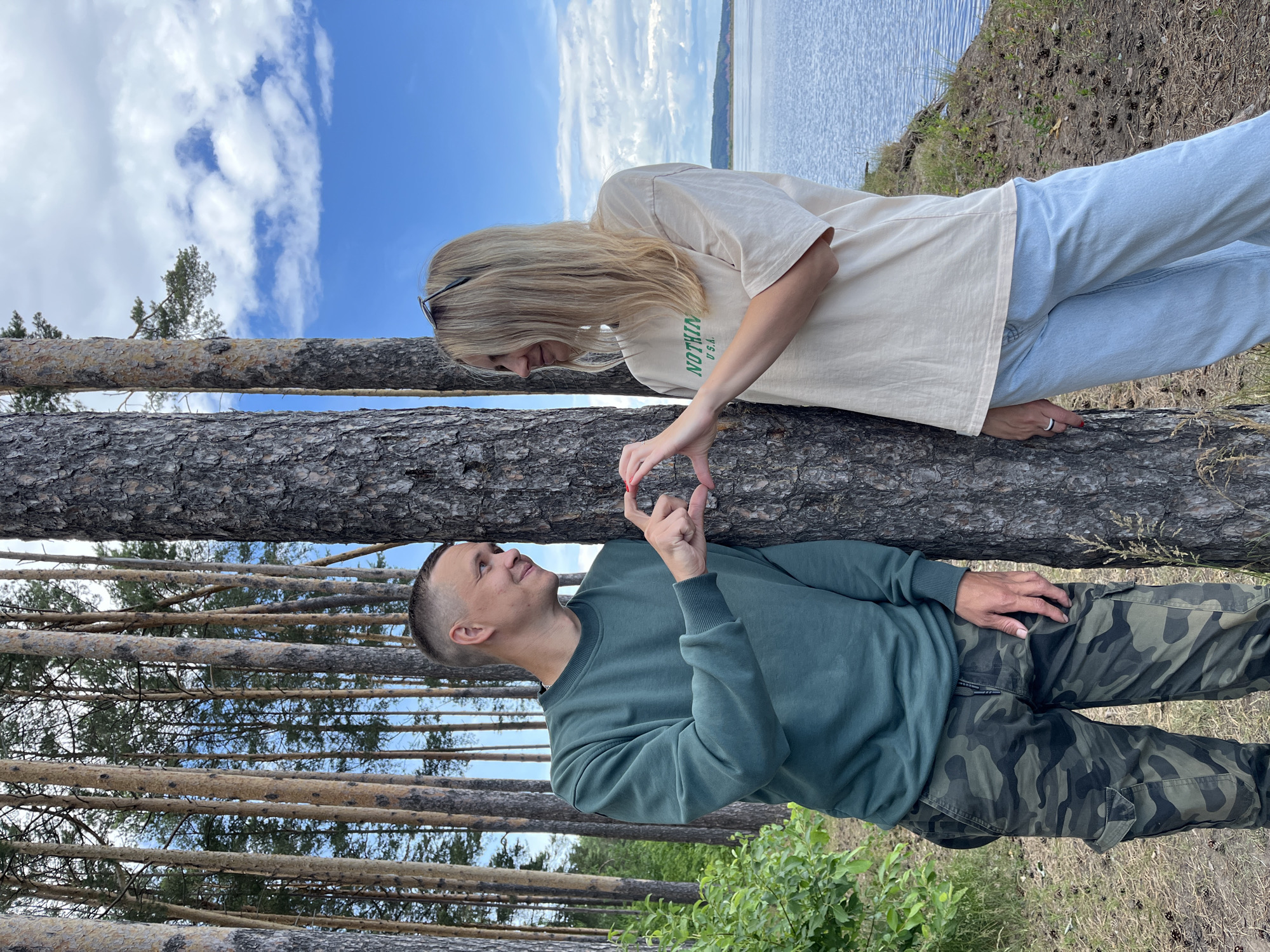 A man and a woman stand next to a pine tree on the bank of the Kama River in Chaikovsky, Russia. The man, wearing a green sweatshirt and camouflage pants, forms a heart shape with his fingers and the woman's. The woman, in a cream-colored t-shirt and light blue jeans, mirrors his gesture. They are both looking at each other and smiling. The background shows a dense forest of pine trees and a bright blue sky with scattered clouds. The river is visible in the distance.