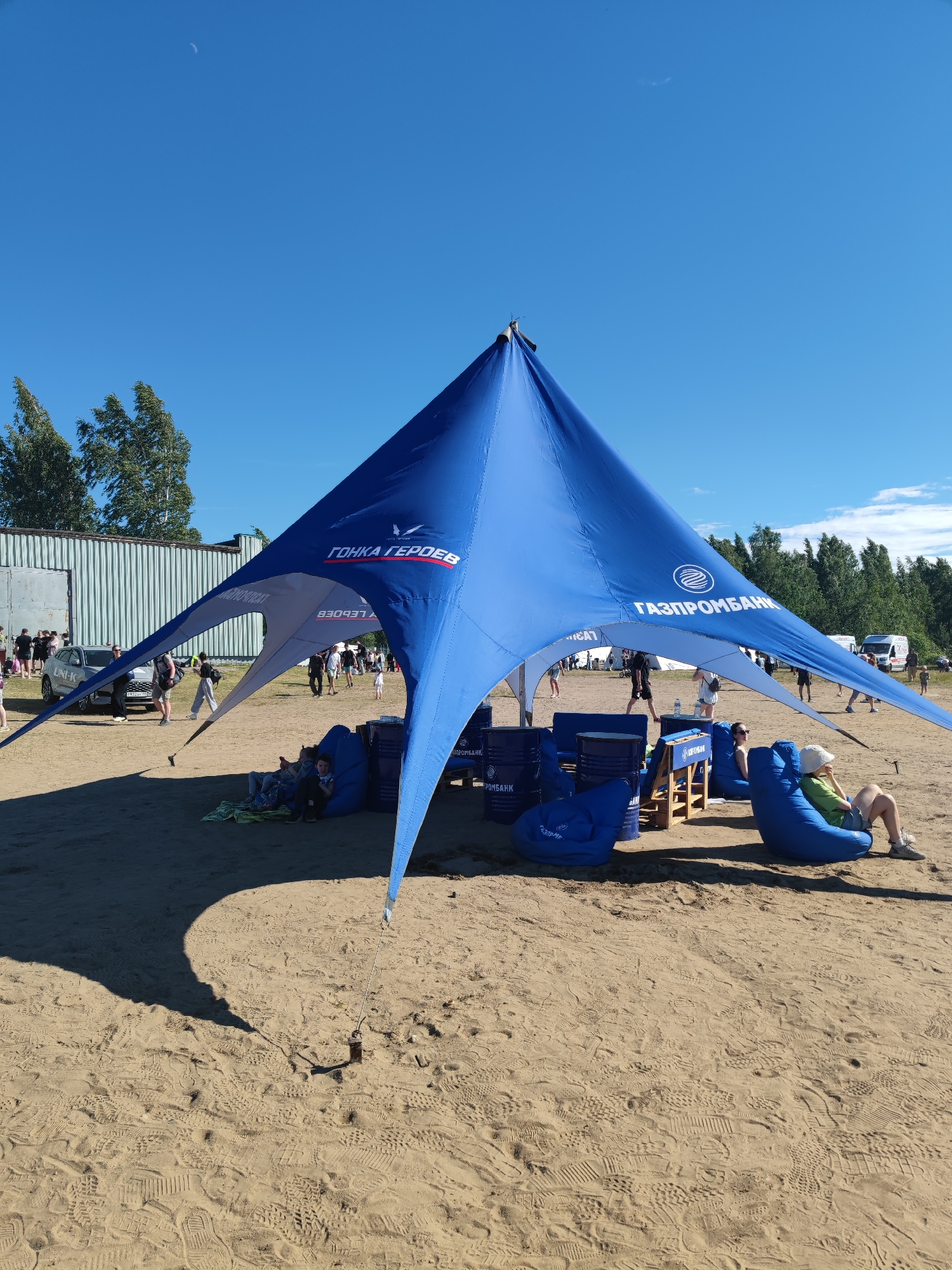 The image captures a bright, sunny day at an outdoor event, likely in the city of Sertolovo, Russia, given the context of the Gazprombank sponsorship visible on the tent. A large, blue, star-shaped tent dominates the foreground, providing shade over a sandy area. Underneath the tent, several blue beanbag chairs are arranged, with two individuals seated on them. One person, wearing a green t-shirt and a white hat, is looking to the right, while another, wearing a white t-shirt and sunglasses, is leaning back. In the background, beyond the tent, a crowd of people is milling around. A silver car with the text "UNI-K" is parked to the left, near a corrugated metal structure. The environment appears to be a sandy, open area, possibly a beach or park, with trees lining the horizon and a clear blue sky above, featuring a crescent moon. The ground is covered with numerous footprints in the sand. The overall scene depicts a casual outdoor gathering, likely a festival or promotional event, under a clear sky.