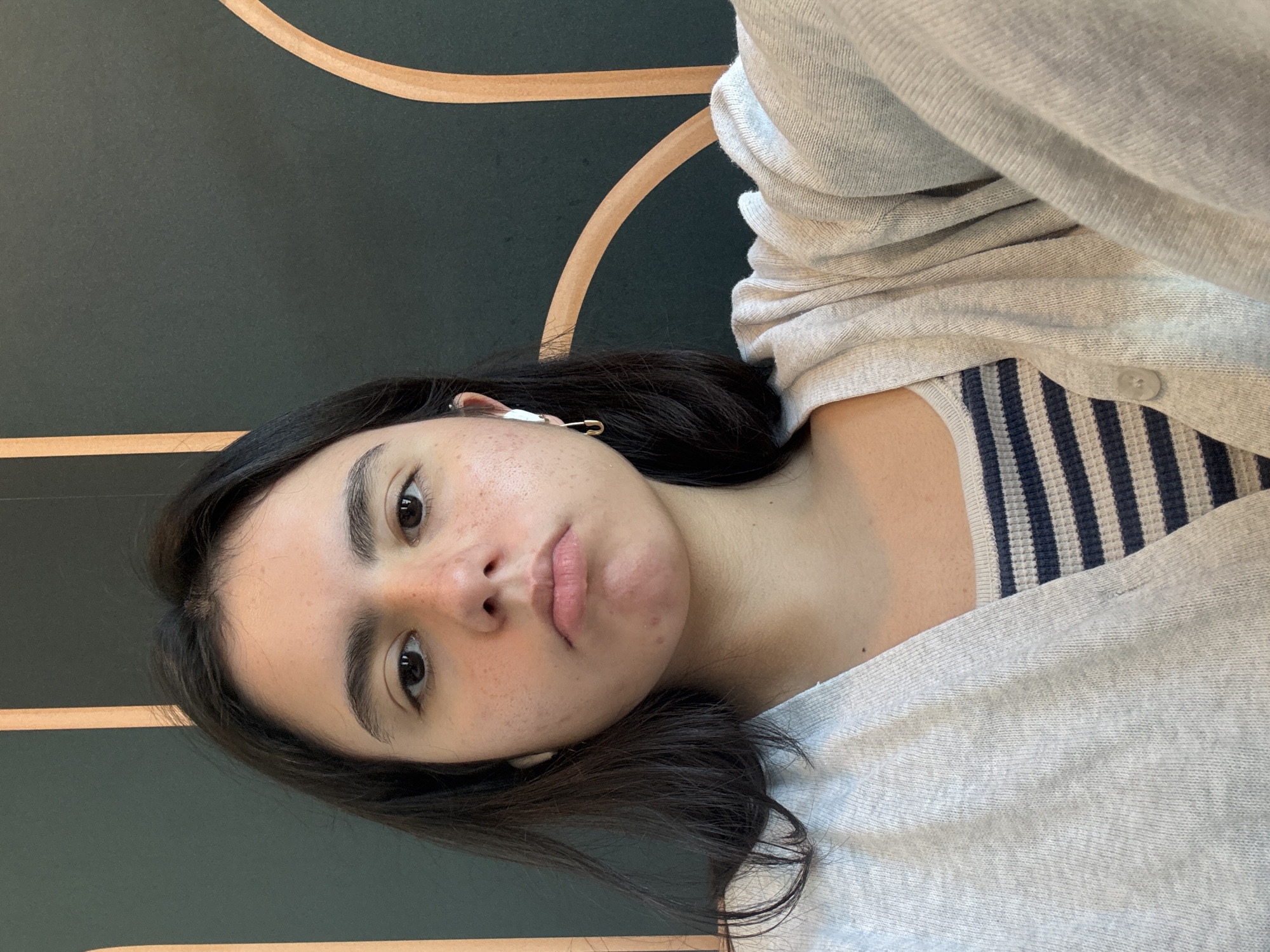 A woman is depicted in a selfie, oriented at an angle. Her hair is dark and she has an earbud in her left ear secured with a safety pin. She wears a striped top and a grey cardigan. The background consists of a dark green wall with curved, light brown lines. There are no clear indicators of the city of Austin.