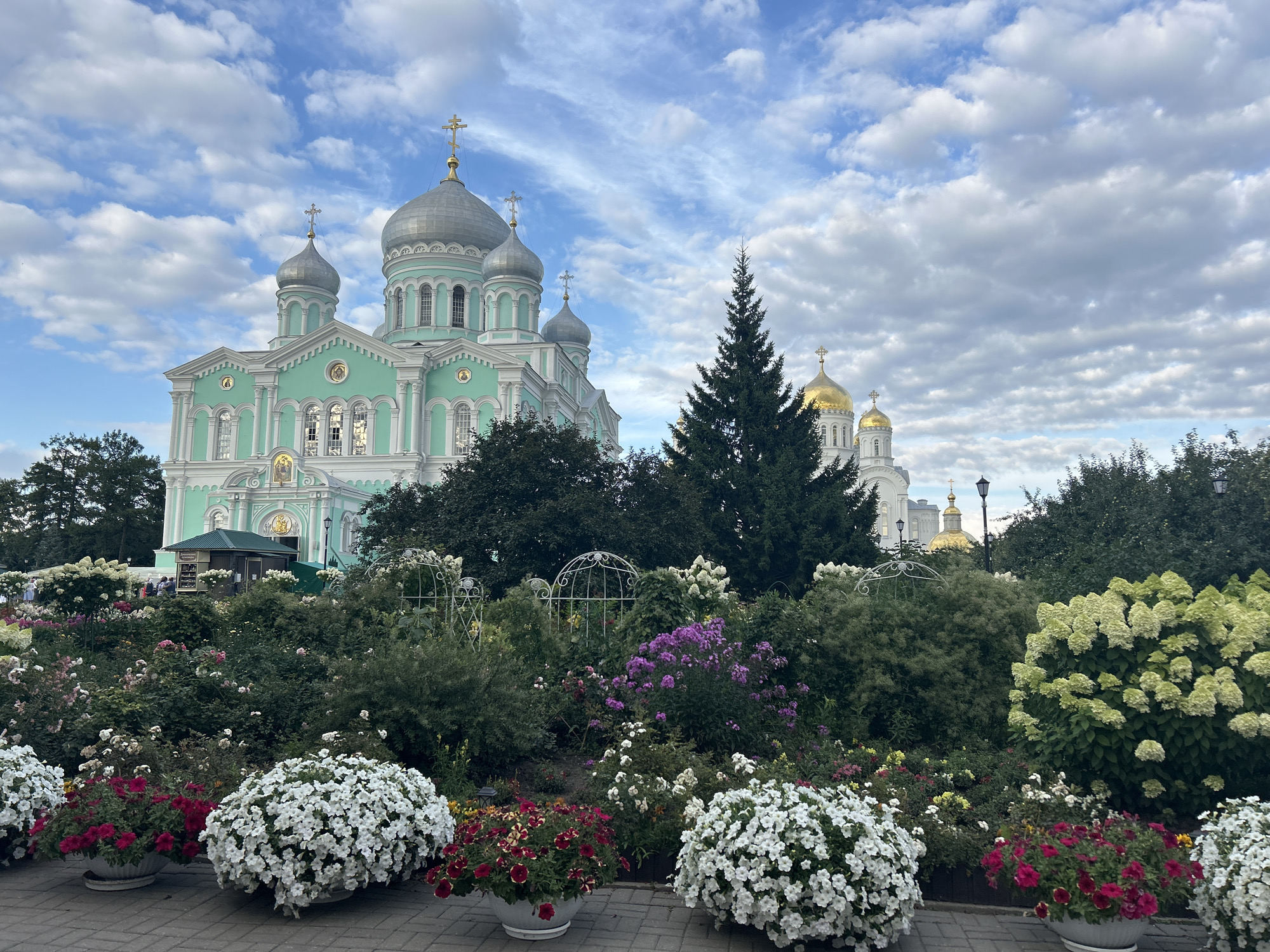 The image captures a view of the Holy Trinity Cathedral in Diveyevo, Russia, under a partially cloudy sky. The cathedral, painted in a mint green and white, features multiple domes topped with crosses. In the foreground, a lush garden is filled with an assortment of blooming flowers, including large clusters of white petunias, pink and red petunias, and large white hydrangea bushes. Several white decorative arches are interspersed within the garden. In the background, behind the garden and trees, another church with golden domes is visible. A small green structure, possibly a kiosk or information booth, is situated in front of the main cathedral. People are subtly visible near this structure. The scene is set on a paved walkway.