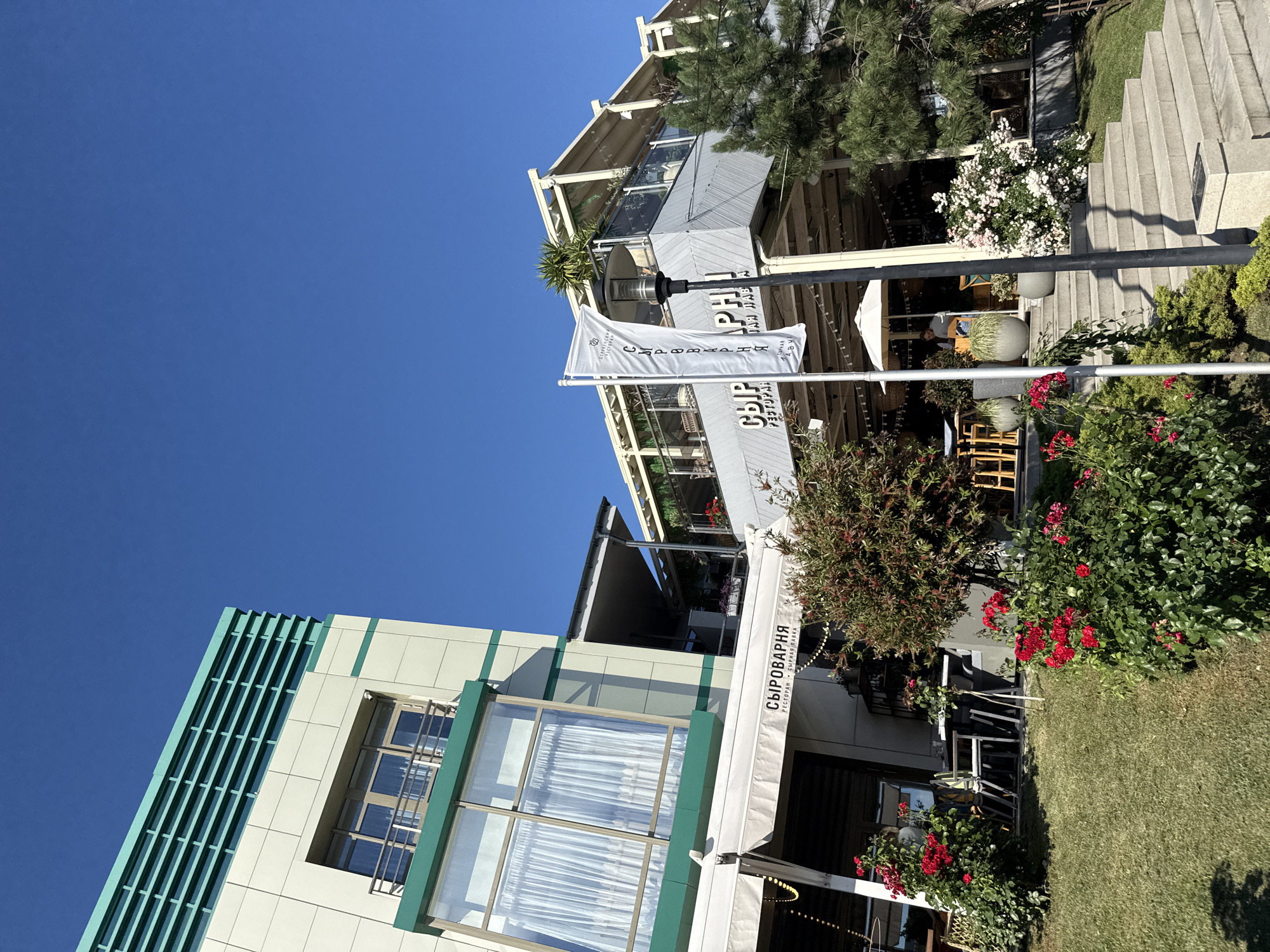 The image shows a multi-story building with a modern design, featuring a light-colored facade and green accents, set against a clear blue sky. The building has large windows and balconies, with awnings extending over outdoor seating areas. Lush green grass and vibrant red roses are visible in the foreground, suggesting a well-maintained outdoor space. A white flag with text is prominently displayed, and a lamppost stands nearby. In the background, several trees and plants contribute to the natural setting. The overall impression is that of a commercial establishment, possibly a restaurant or cafe, in a pleasant outdoor environment. This scene is located in Sochi, Russia, likely a coastal city known for its pleasant climate and numerous recreational facilities.
