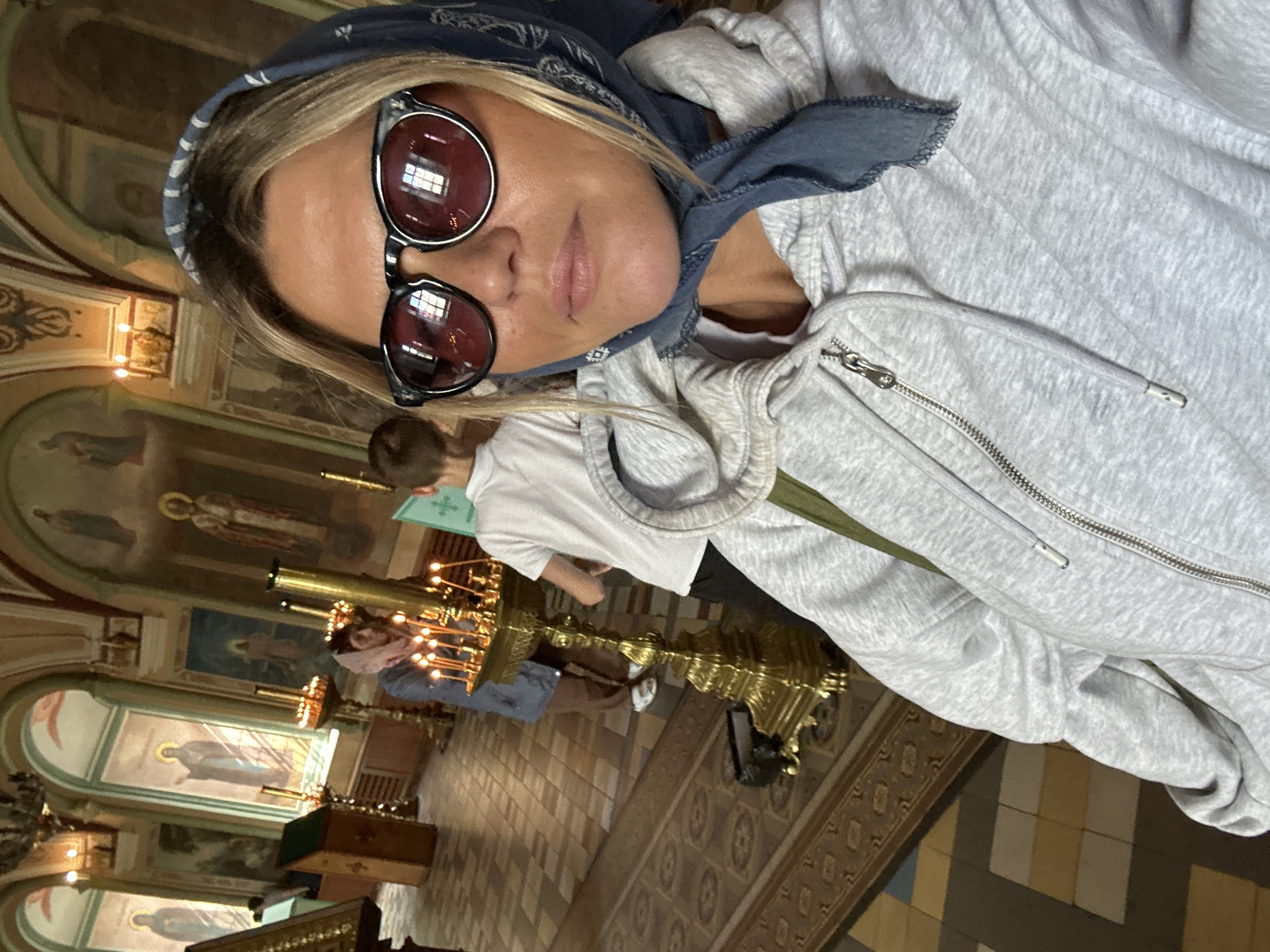 A woman wearing sunglasses and a blue headscarf takes a selfie in a church. She is wearing a light gray hooded sweatshirt. In the background, there are religious paintings on the walls, candles on a golden candelabra, and other people in the church. The floor is tiled in a checkered pattern. The scene appears to be inside the Trinity Lavra of St. Sergius in Sergiyev Posad, Russia, given the architectural style and religious iconography.