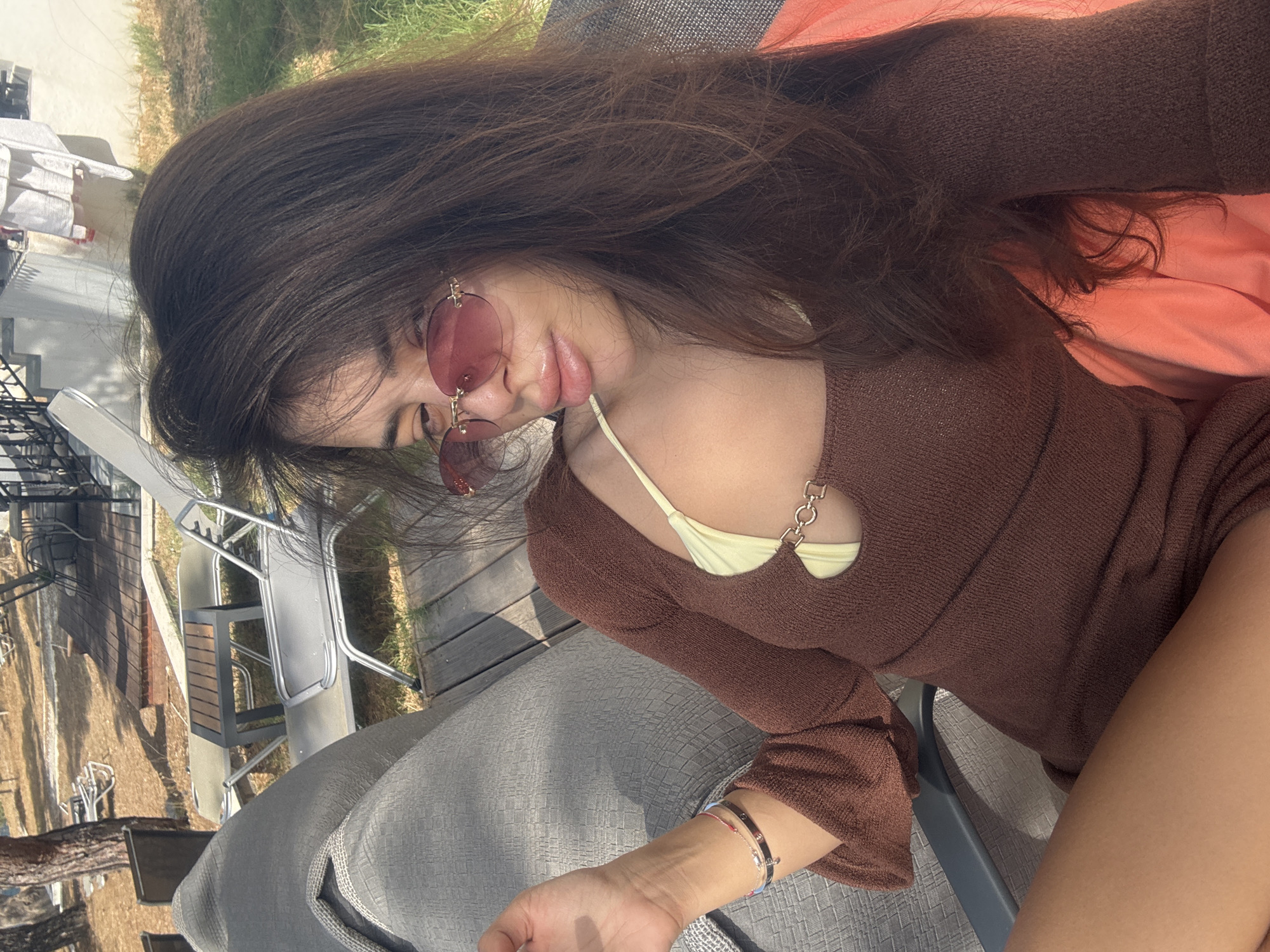 A woman with long, dark brown hair and pink-tinted oval sunglasses is posing for a selfie outdoors. She is wearing a brown, long-sleeved, cutout dress with a light yellow bikini top underneath. She has bracelets on her left wrist. The background shows a wooden deck with chairs and tables, and vegetation. The scene is likely a resort or outdoor seating area in Greece.