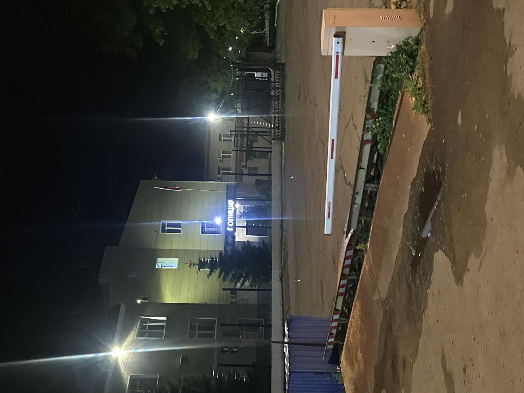 The image depicts a street scene at night, likely in Birobidzhan, Russia. A white and red striped barrier arm is prominently featured in the foreground, suggesting a controlled access point such as a parking lot or a private road. Behind the barrier, a modern building with multiple windows is visible, illuminated by artificial lights, casting a yellowish glow. One section of the building has illuminated text that reads "ПОЛИЦИЯ," which translates to "POLICE" in Russian. Trees with green foliage are interspersed around the building, some lit by streetlights, creating a contrast between the darkness of the night and the illuminated elements. The ground appears to be asphalt, with some patches of wetness or oil stains, indicating it might have rained recently or there is some residue on the road. The overall atmosphere is quiet and still, characteristic of a late-night urban or semi-urban environment. The time of day is clearly night due to the artificial lighting and the dark sky. There are no people visible in the frame, emphasizing the stillness of the scene.