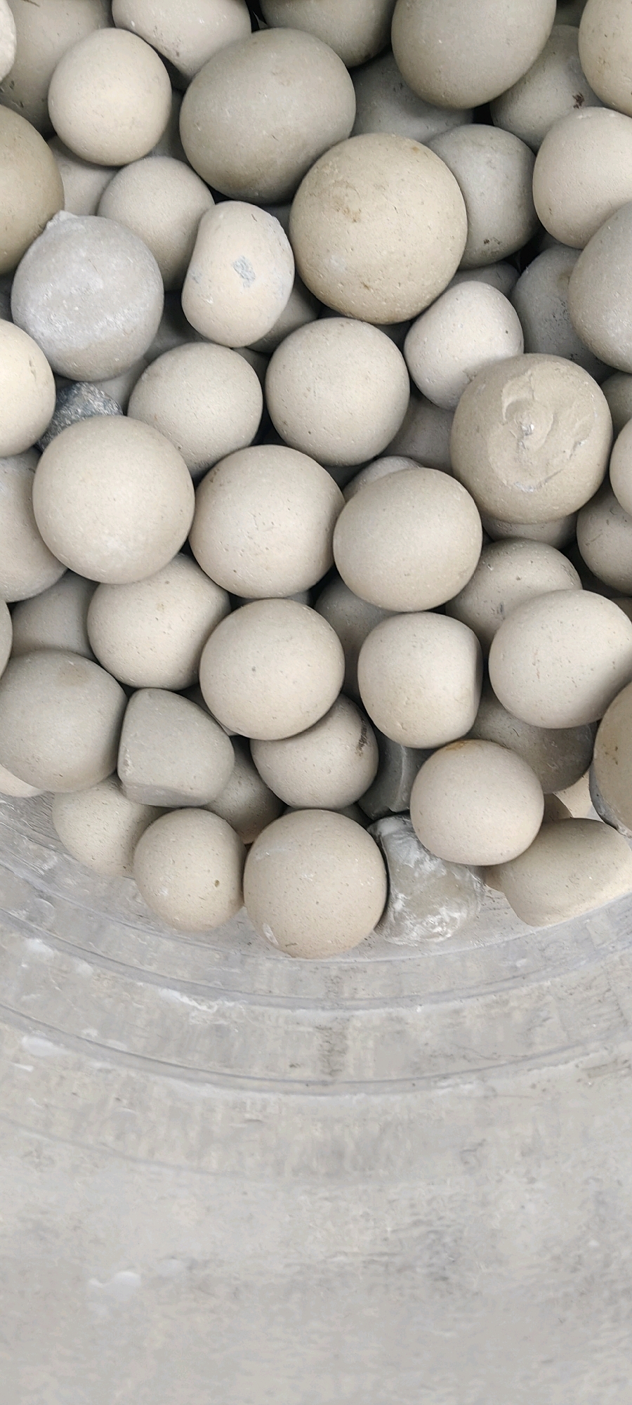 The image displays a close-up, overhead view of a collection of small, round, pale beige stones or pebbles filling a container. The stones are of various sizes, with some appearing perfectly spherical while others are slightly irregular or flattened. The surface texture of the stones is matte and slightly dusty. The container's rim, made of a ribbed, light gray material, is visible at the bottom of the frame. The background is blurred, suggesting a shallow depth of field, and appears to be a continuation of the container's material or a similar surface. There are no people or discernable actions taking place. The scene is static and focuses solely on the inanimate objects. The location is not specified within the image itself, and therefore cannot be determined from the visual information provided.