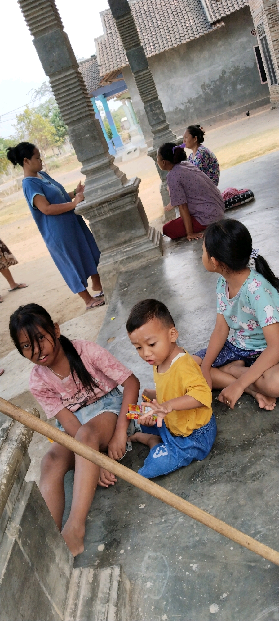 In Tanjung Ratu Ilir, Indonesia, two young children are seated on a concrete floor. A girl with dark hair and bangs, wearing a pink t-shirt and denim shorts, is looking towards the camera with an open mouth. Beside her, a boy in a yellow t-shirt and blue pants is holding a pack of candy, looking down at it. Another child, a girl with dark hair in a ponytail wearing a light blue t-shirt with cartoon characters, is partially visible to the right, also seated on the floor. In the background, two adult women are seated. One woman in a purple floral top and maroon pants is looking away, while another woman in a blue dress is standing and gesturing with her hands. The environment appears to be an outdoor covered area with concrete pillars and a tiled roof structure visible. The ground is dusty and has some scattered debris.