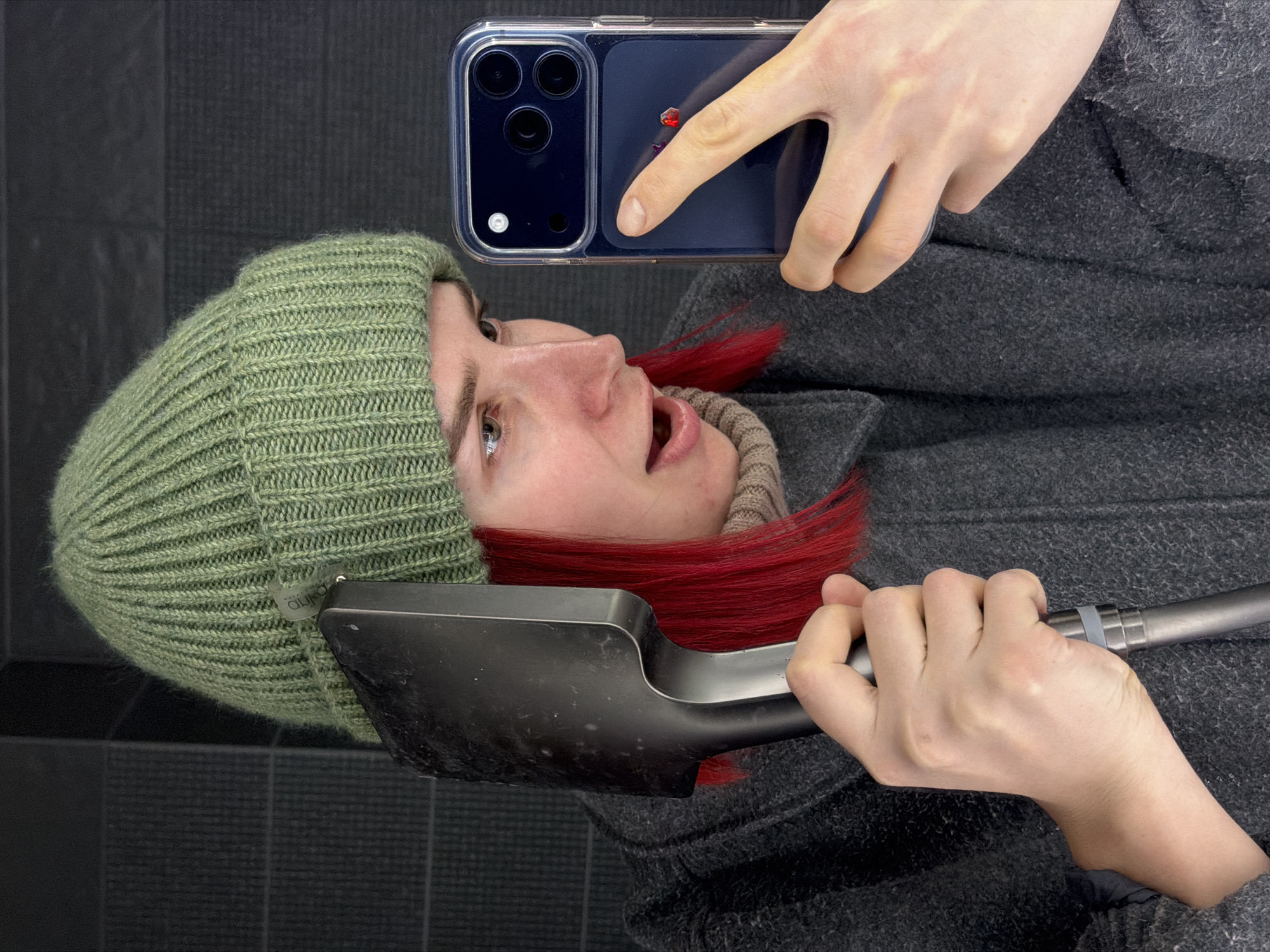 This selfie captures a person, likely female, from the chest up, in what appears to be an indoor or enclosed setting in Chaikovsky, Russia. The image is oriented sideways, with the subject's head to the left and body to the right, but the action suggests an upright stance.
The person has fair skin, visible red hair, and light-colored eyes. They are dressed for cold weather, wearing a ribbed, light olive green knitted beanie hat and a dark gray, textured coat. A beige, high-neck sweater is visible beneath the coat.
In their right hand, they hold a dark blue smartphone in a clear case, with the multi-lens camera module prominently displayed. A finger is pressing a button on the side of the phone, indicating they are actively taking the selfie. Their left hand firmly grips the handle of a dark gray or black compact shovel, possibly a snow or camping shovel, which extends across the lower portion of the frame.
The person's facial expression conveys a sense of slight surprise or playfulness, with their mouth slightly open and their eyes looking upwards towards the camera lens. The background is a dark, uniform surface, possibly a tiled wall or vertical panels, providing a neutral backdrop. A white tag is visible on the beanie, but its text is illegible.