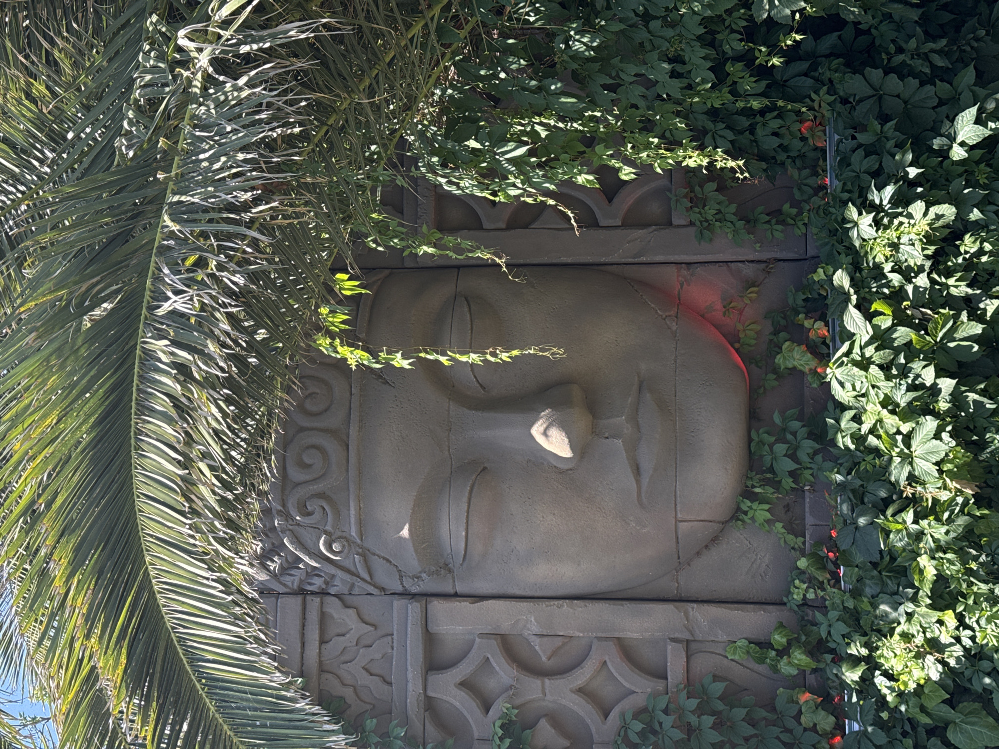 The image depicts a large, stone relief of a serene face, possibly a Buddha or a deity, embedded in a wall. The relief is adorned with intricate patterns on either side of the face. Lush green foliage, including palm fronds and vines, partially obscures the stone structure, creating a natural, somewhat overgrown aesthetic. Red accents are visible among the foliage, possibly flowers or decorative lights. The overall scene suggests an outdoor setting, possibly a garden or a decorative wall feature in a public space. The lighting indicates it is daytime, with sunlight filtering through the leaves. The location of this artwork is in Asprovalta, Greece.